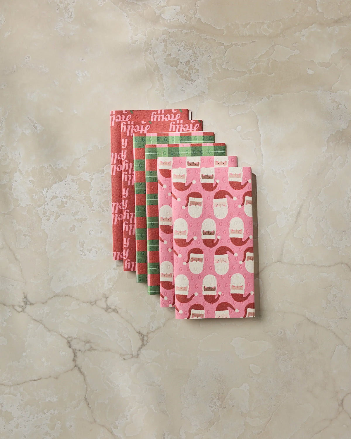 Four folded pieces of Geometry’s Holly Jolly Santa holiday-themed wrapping paper, featuring vibrant red, green, and pink patterns with festive text and Santa faces, are arranged on a light marble surface—perfect for pairing with eco-friendly Geometry kitchen towels to create sustainable and cheerful gift presentations.