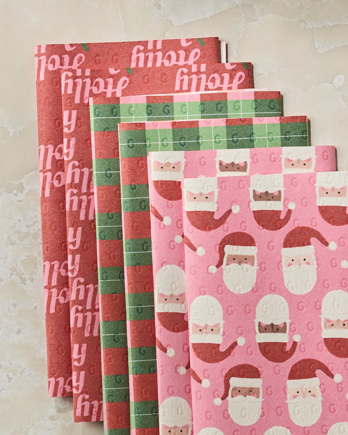 Bring holiday cheer to your table with the Holly Jolly Santa cocktail napkin set from Geometry. This five-piece collection features joyful holiday patterns—an all-red napkin with “hip hip hooray” lettering, a classic green and red plaid, plus two playful pink designs adorned with Santa Claus faces. Designed to look like reusable paper towels, they’re displayed here on a light marble surface, adding a festive touch to any gathering.