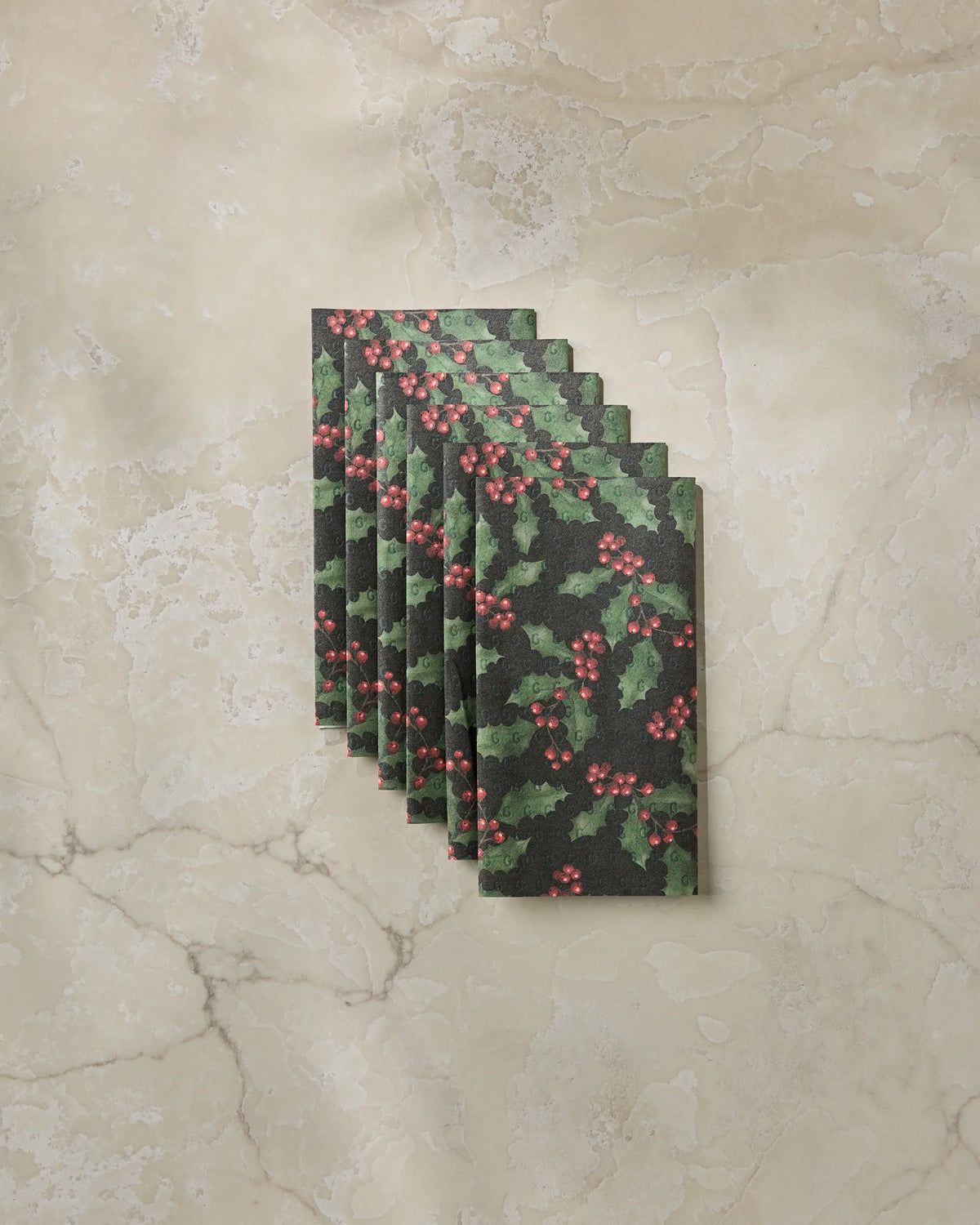 Six rectangular Holly Night napkins by Geometry, featuring a festive holly and berry pattern, are neatly stacked on a beige marble surface—providing an elegant and eco-friendly alternative to disposable paper towels.