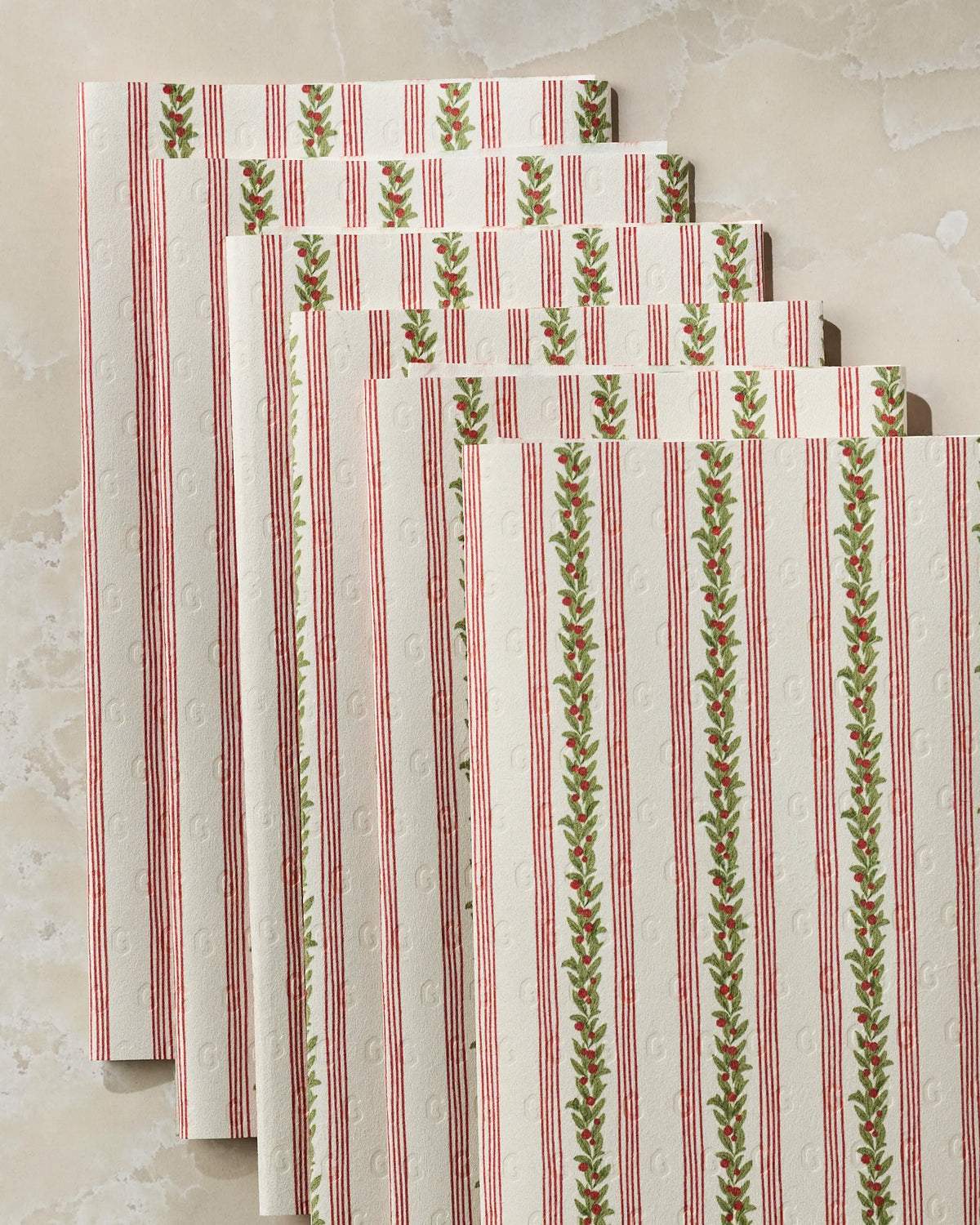 Five sheets of Geometry’s Holly Vine reusable paper towels, featuring cream-colored fabric with red vertical stripes and green leafy patterns, are neatly stacked on a light marble surface.