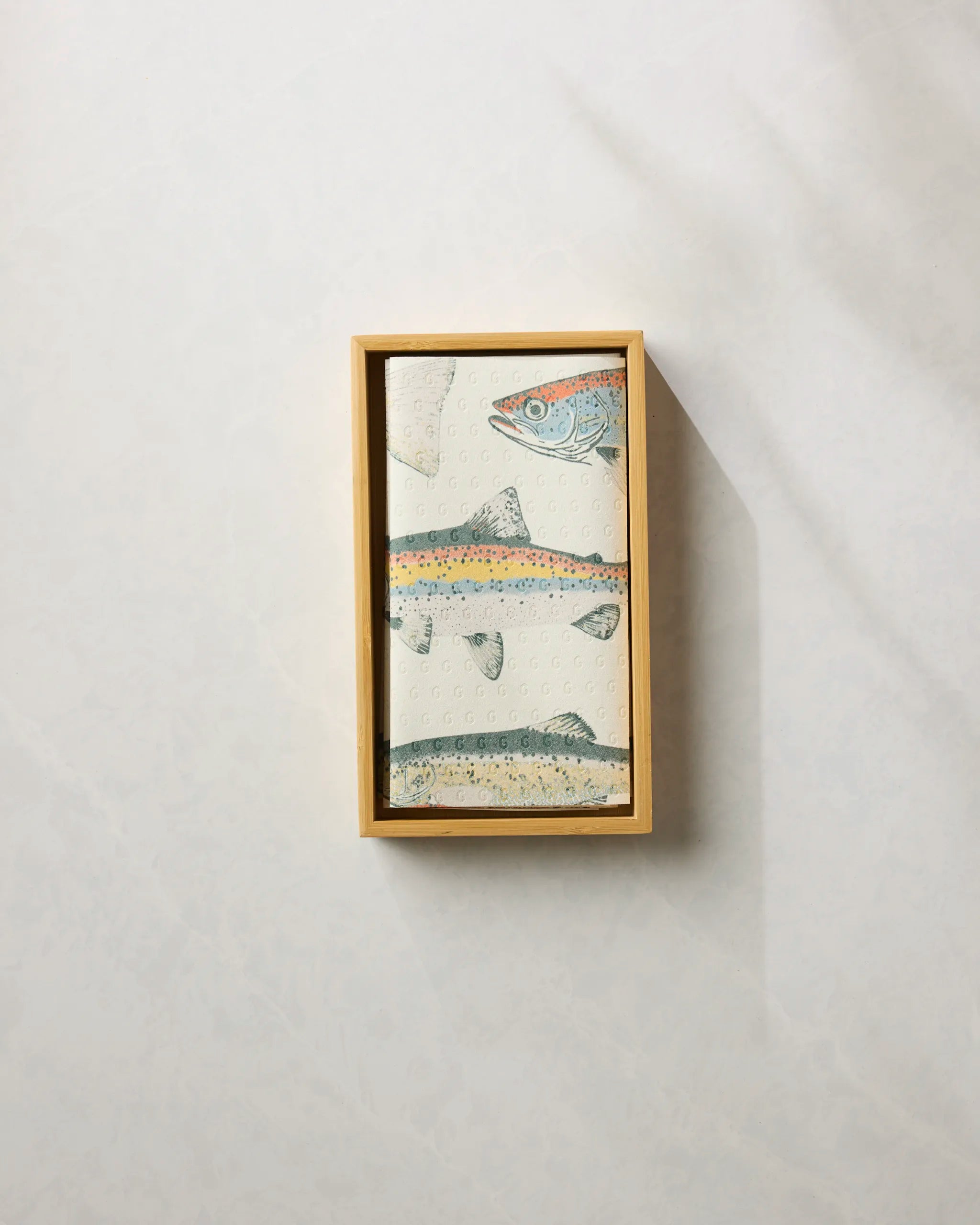 A neatly folded Geometry "Hooked on Fishing" reusable paper towel featuring an illustrated fish design sits in a rectangular wooden box against a light, textured background.