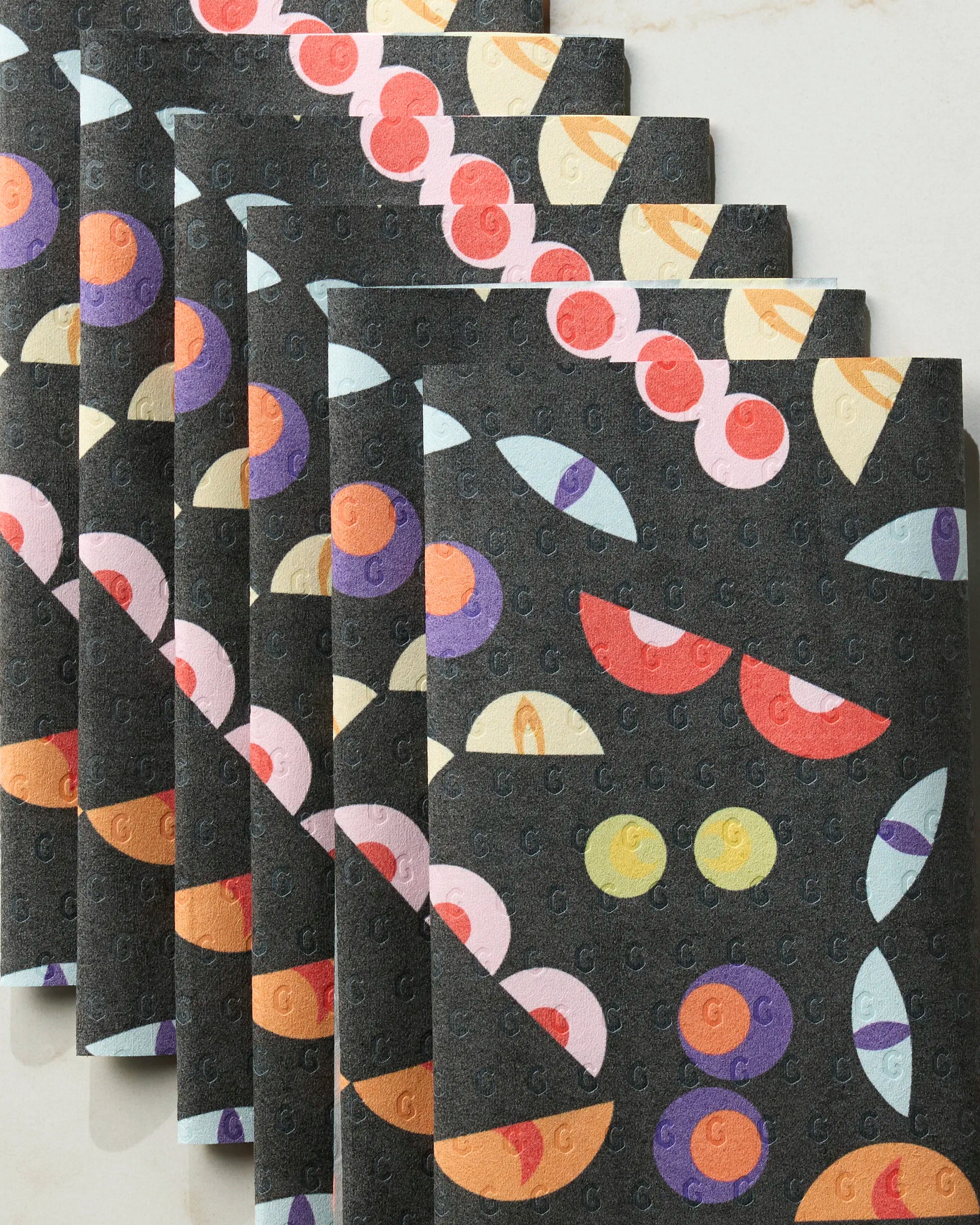 Six folded Geometry “I See You” reusable paper towels featuring colorful abstract fruit patterns on black are fanned out on white marble, adding an eco-friendly touch.