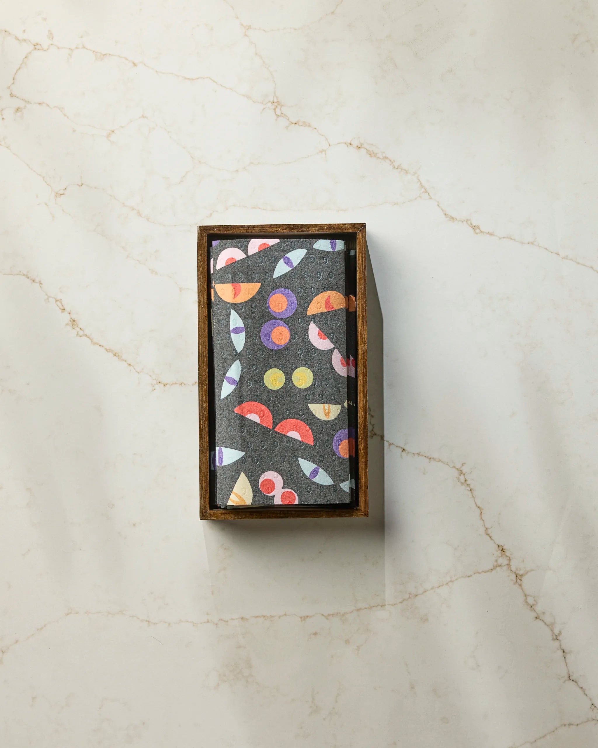 The Geometry "I See You" rectangular box has a black lid with a vibrant abstract pattern of circles, semicircles, and lines, designed for eco-friendly paper towels. It’s shown on a white marble surface with light brown veins.