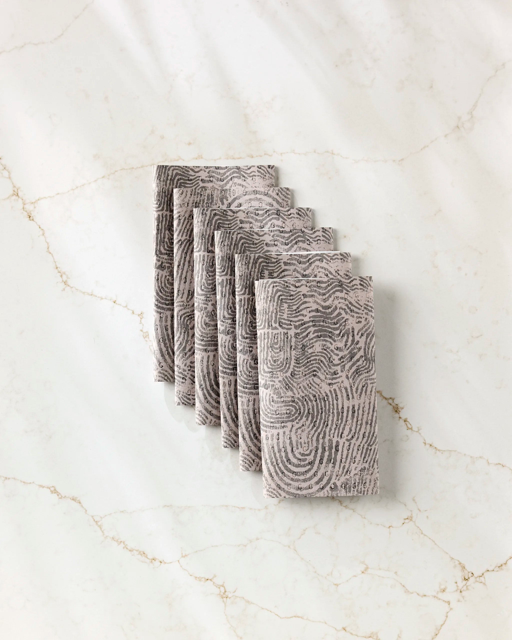 Six gray "Imprint" cloth napkins by Studio Jeannot, featuring a fingerprint-like swirl pattern and ideal as reusable paper towels, are neatly arranged on white marble with light brown veining.
