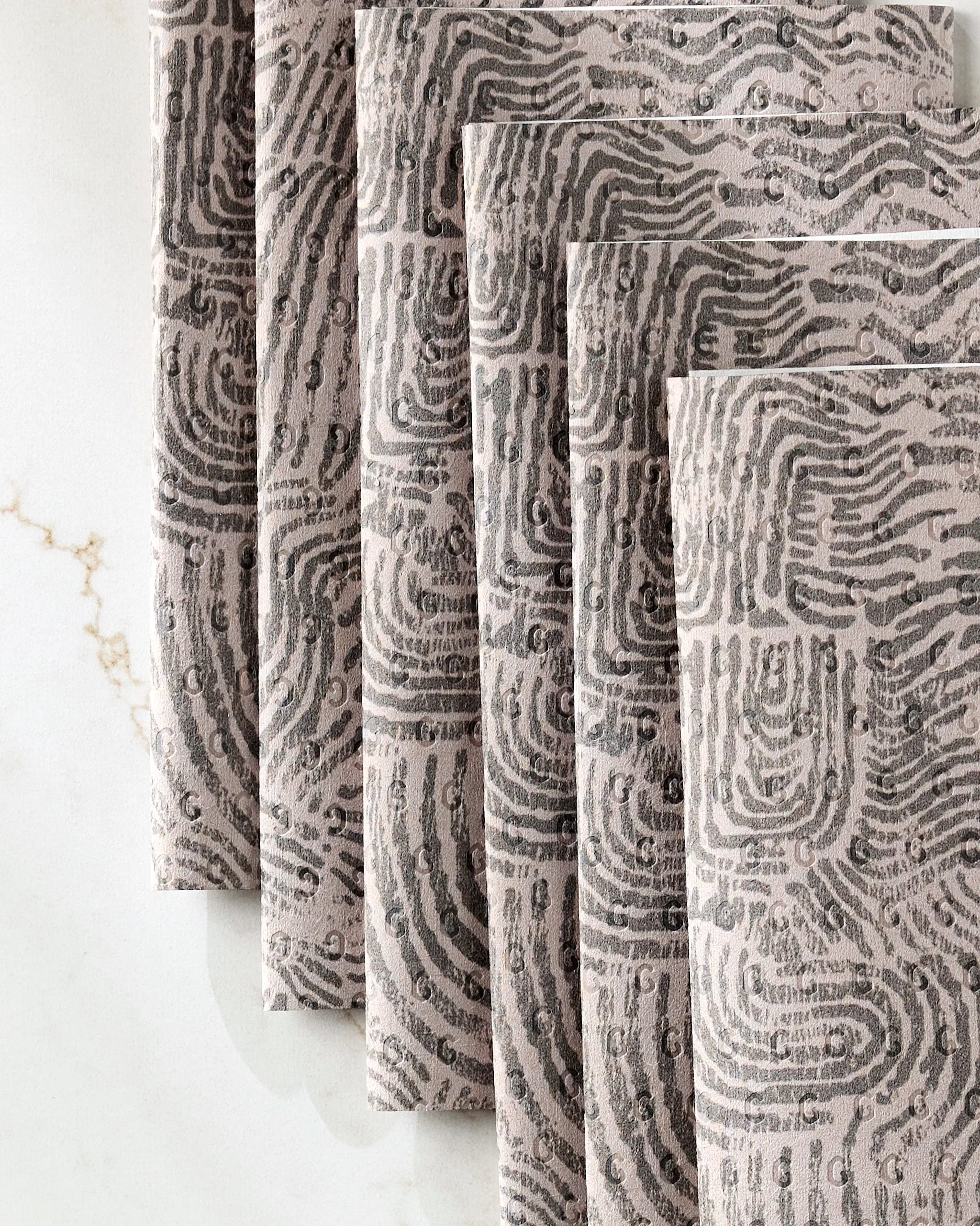 Five “Imprint” cloth napkins by Studio Jeannot, featuring a gray and black abstract fingerprint pattern, are elegantly stacked on a white marble surface with subtle gold veining—a chic alternative to disposable paper towels.