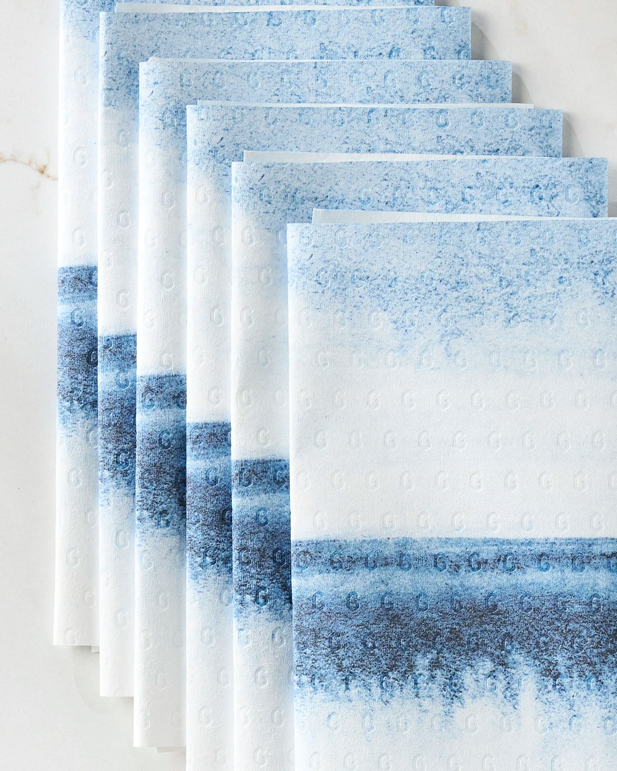 Five white "In The Distance" napkins by GEOMETRY, featuring a blue gradient landscape and an embossed G, are fanned on a white surface—an elegant, eco-friendly kitchen towel choice.