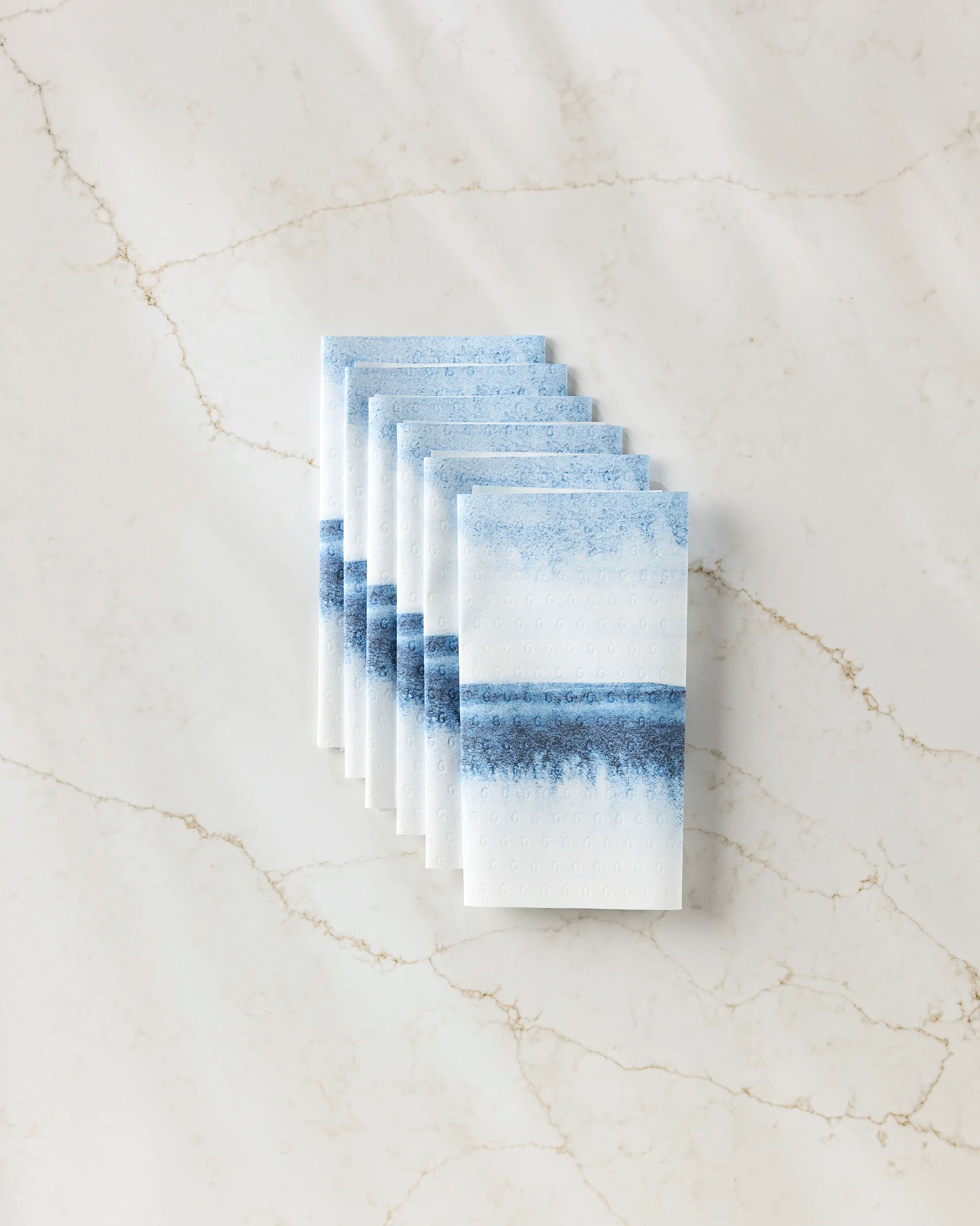 Eight stacked "In The Distance" kitchen towels by GEOMETRY, featuring a blue watercolor stripe design, are arranged neatly on beige-veined marble. Eco-friendly and stylish for any kitchen.