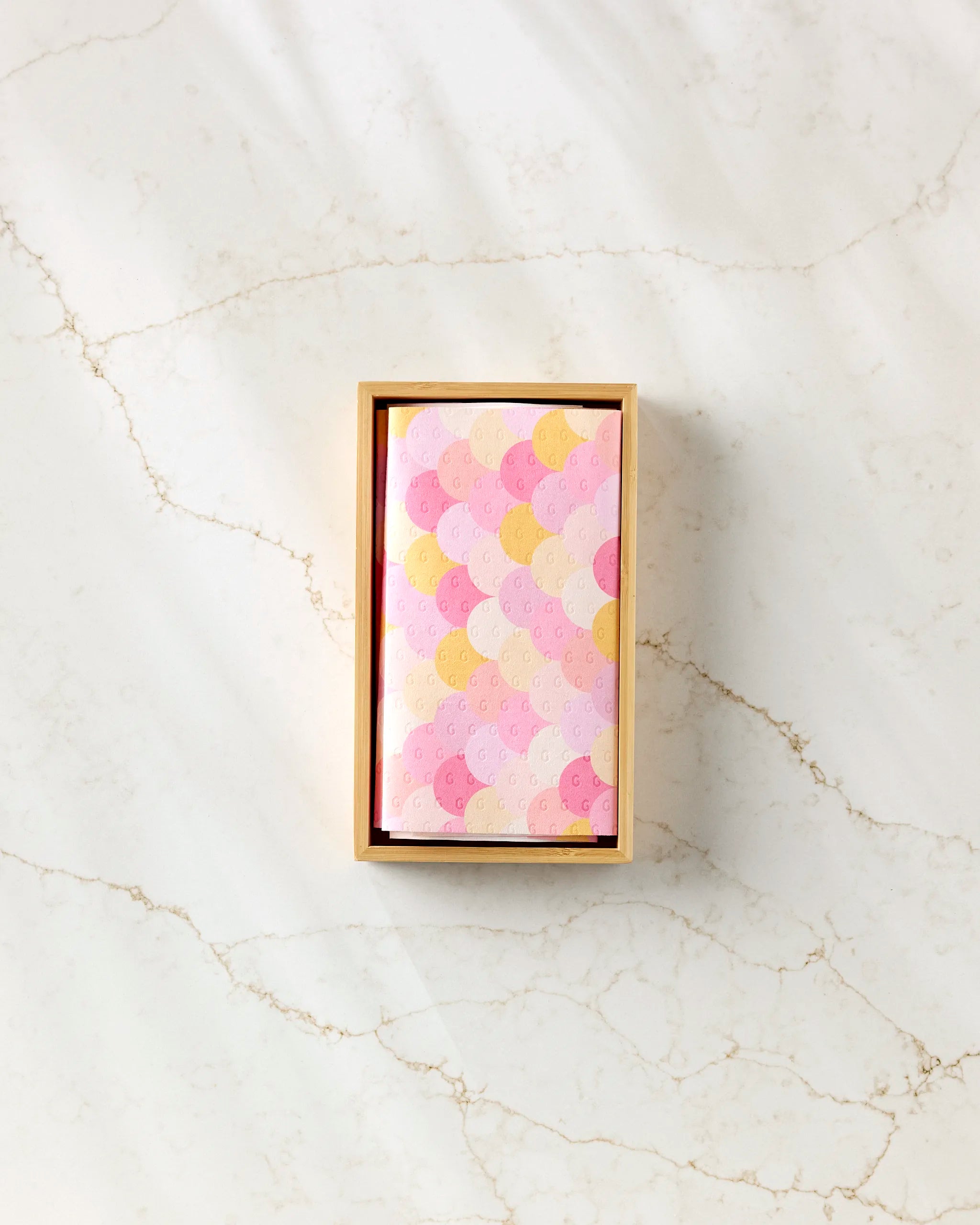 A rectangular box of GEOMETRY's Inherit eco-friendly kitchen towels, featuring a pastel scallop design with pink, yellow, and light purple arcs, rests on a light marble surface with soft shadows.