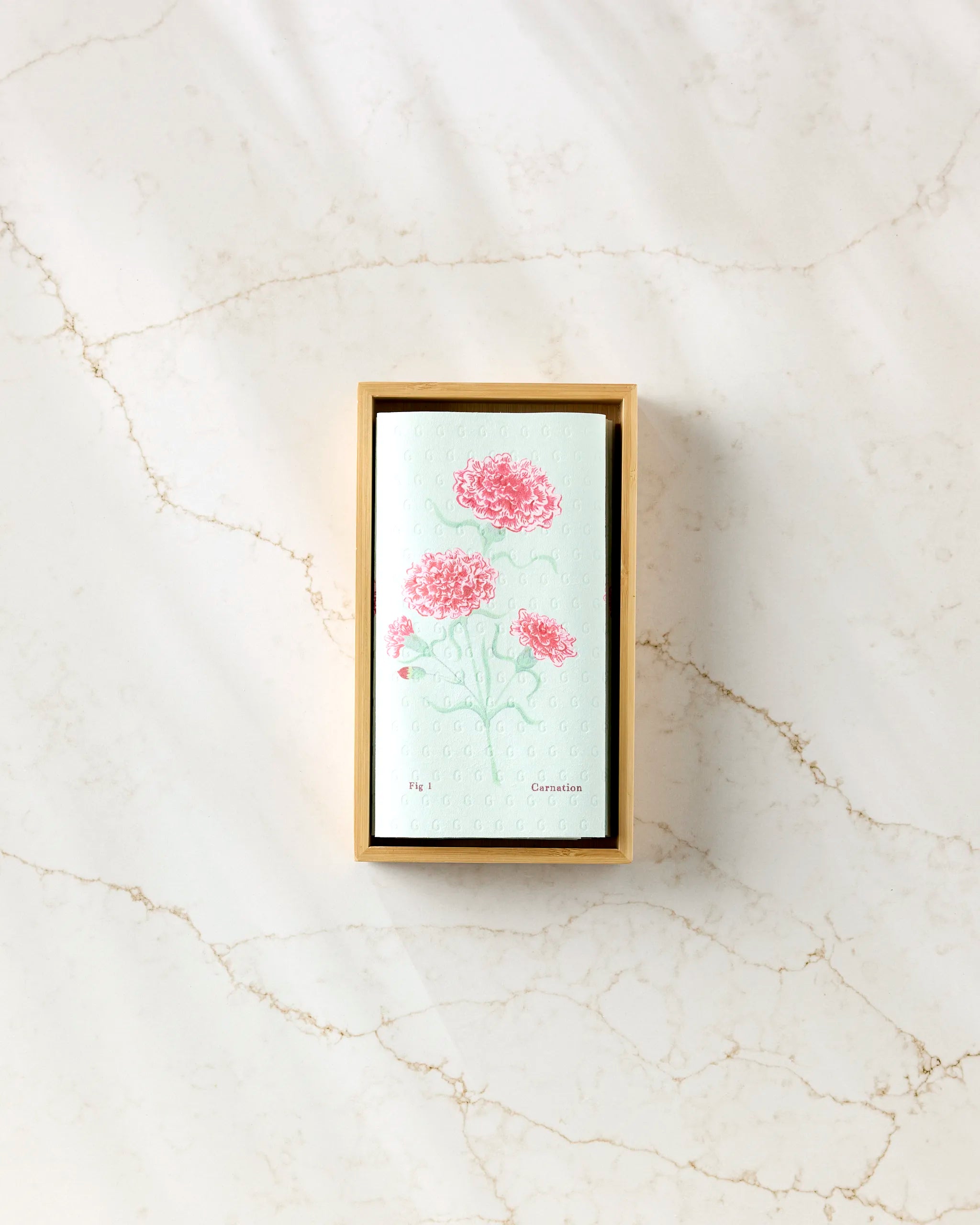 The Linda Fox "January Carnation" eco-friendly paper towels come in a rectangular box adorned with pink carnation florals on a light mint background, displayed on a marbled white surface with soft shadows.