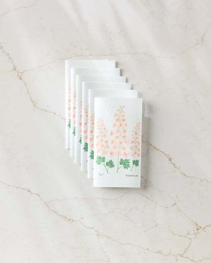 Seven white July Delphinium eco-friendly paper towels by Linda Fox, featuring pink and green floral designs, are neatly stacked in a fan arrangement on a light marble surface.