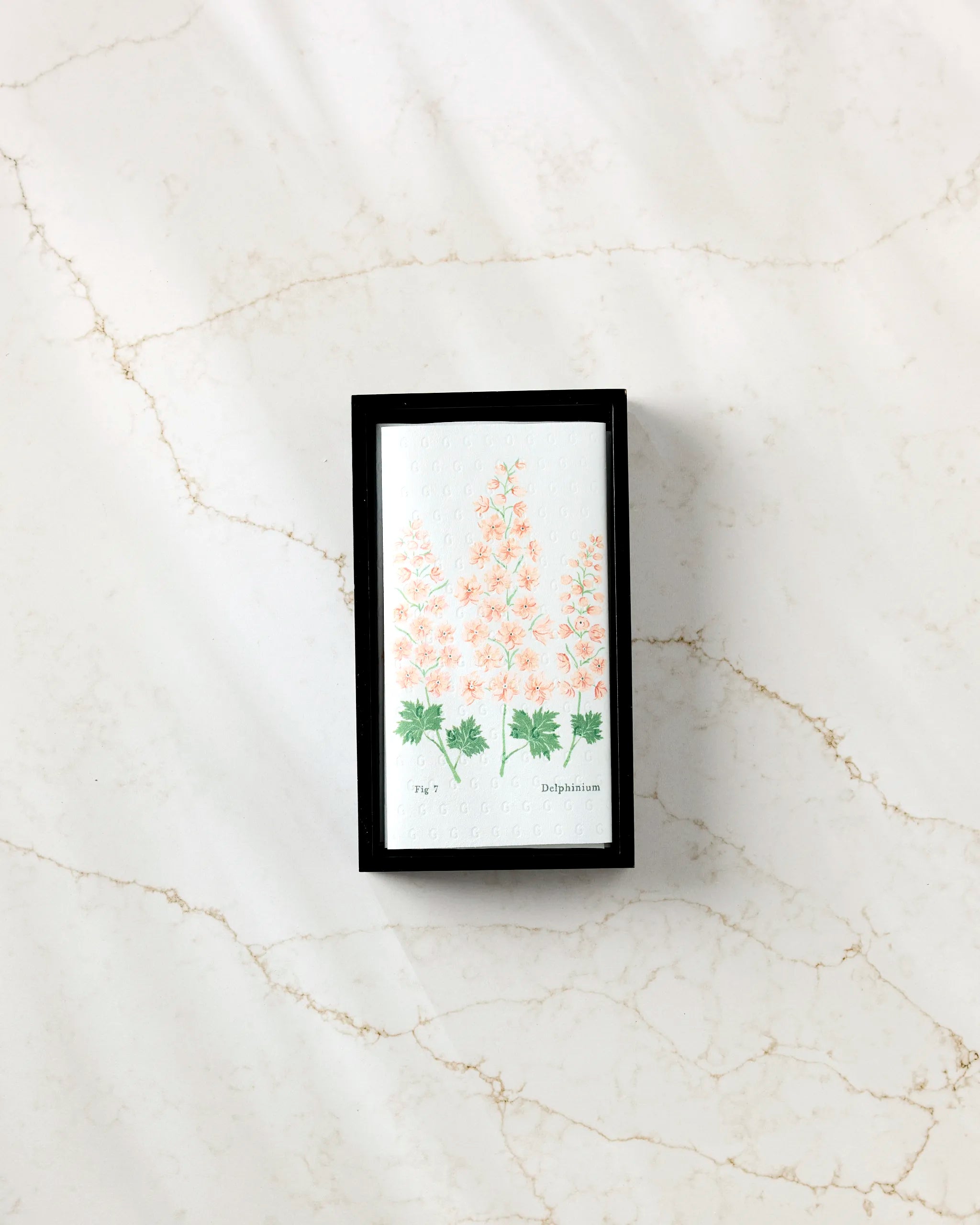 The Linda Fox July Delphinium is a small rectangular box for eco-friendly paper towels, adorned with pale pink flower and green leaf illustrations on the lid, shown on a white marble surface with light brown veining.