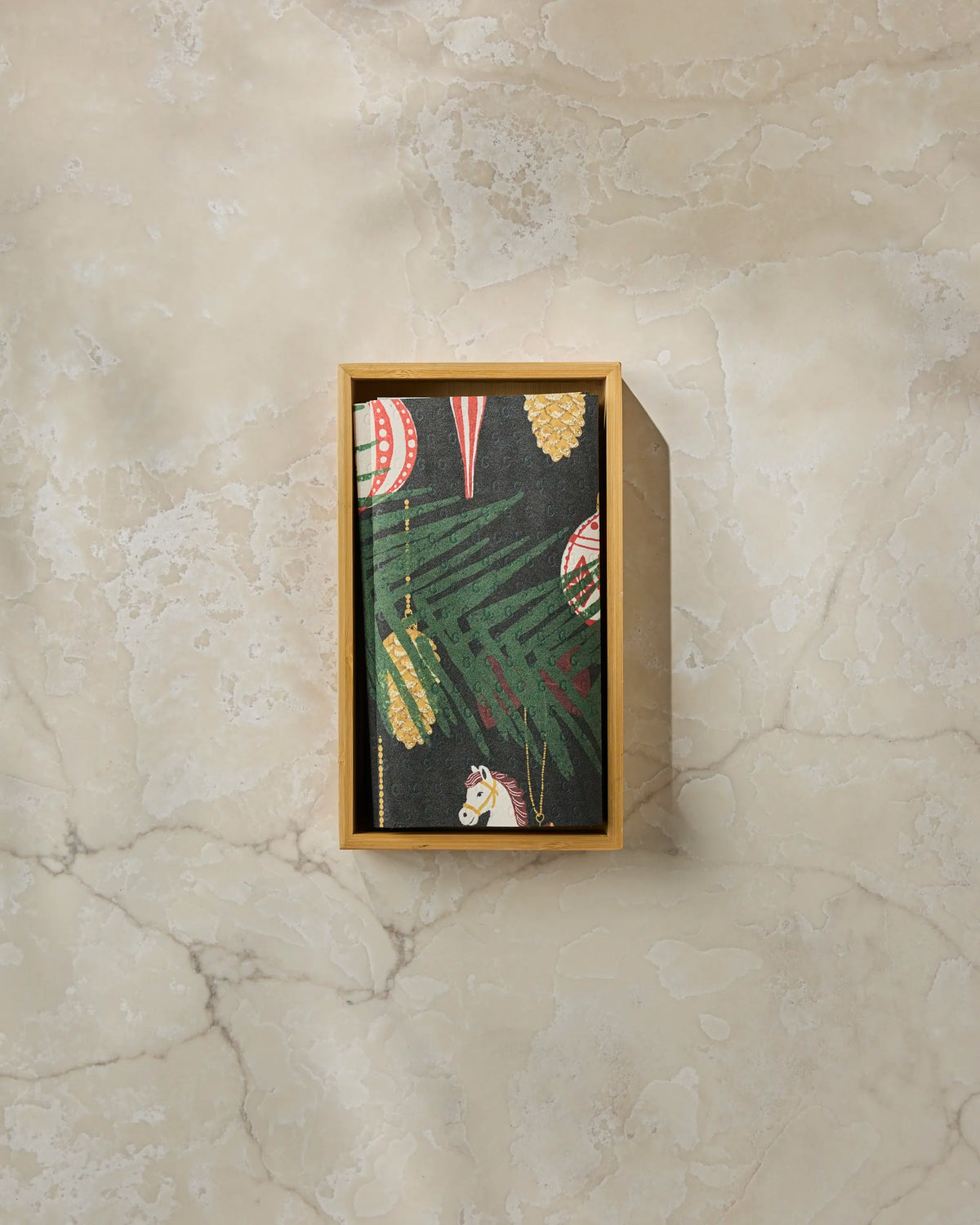 A closed Geometry Keepsake notebook, featuring a dark tropical leaf and ornament pattern, rests inside a wooden box on a beige marble surface and is paired with reusable paper towels for an eco-friendly touch.