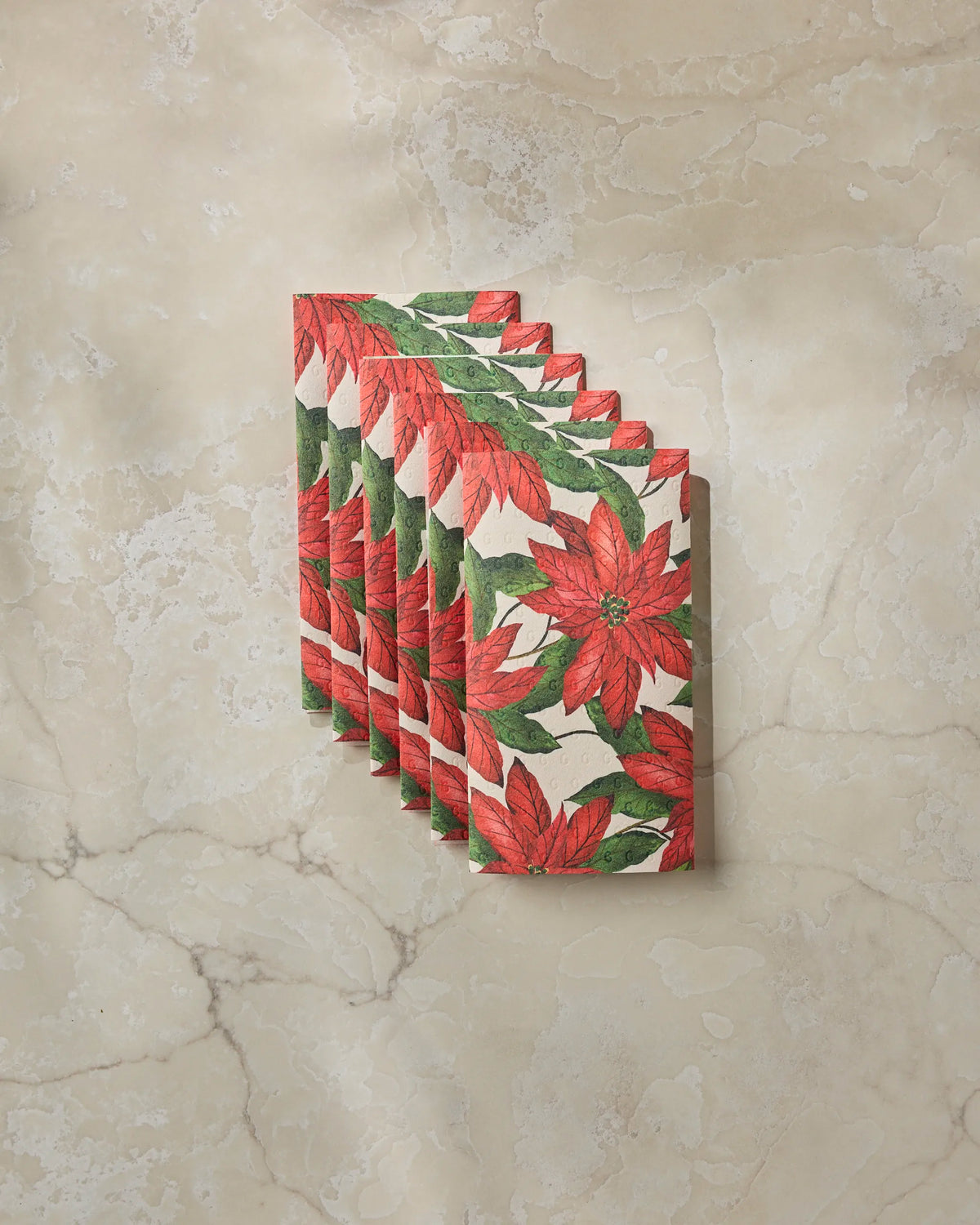 Seven Lauri cloth napkins by Geometry, featuring red poinsettias and green leaves, are fanned out on a beige marble surface—providing an environmentally friendly alternative to disposable options.
