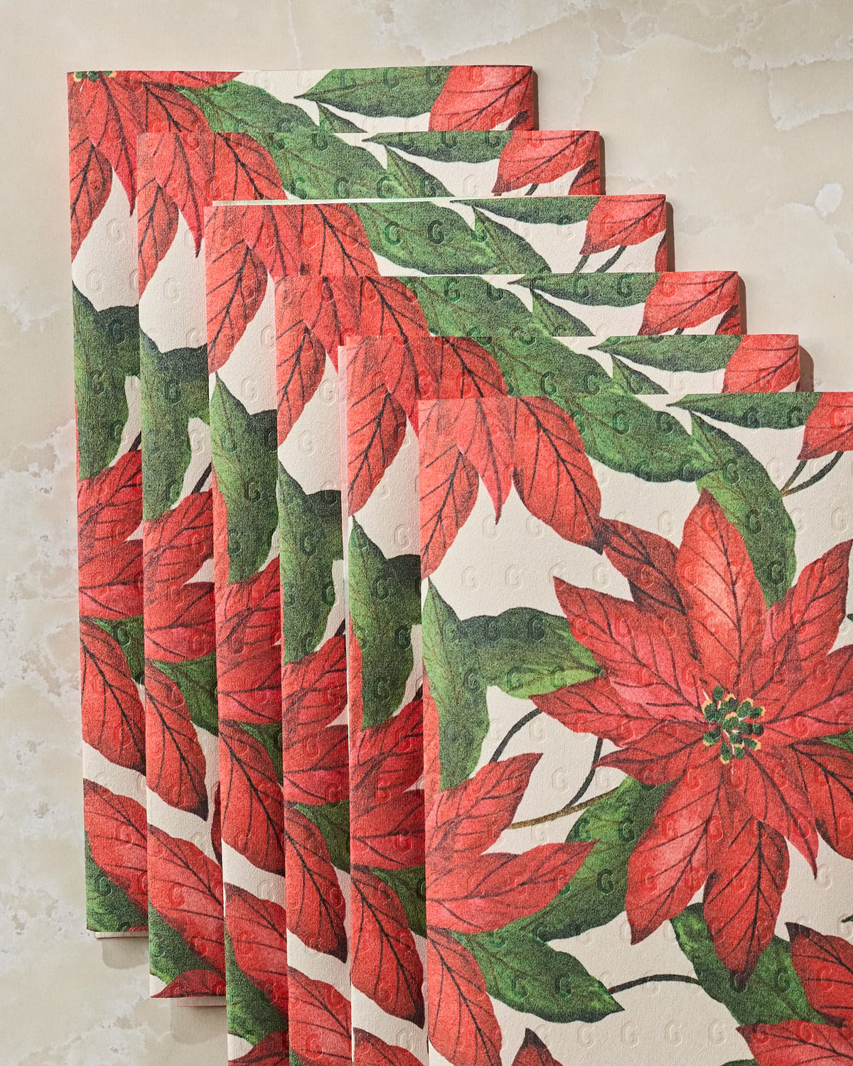 Five Lauri napkins by Geometry, made from post-consumer recycled materials and featuring red poinsettia flowers with green leaves, are arranged in a fanned-out pattern on a beige marbled surface. These environmentally friendly alternatives add festive charm while helping reduce waste.