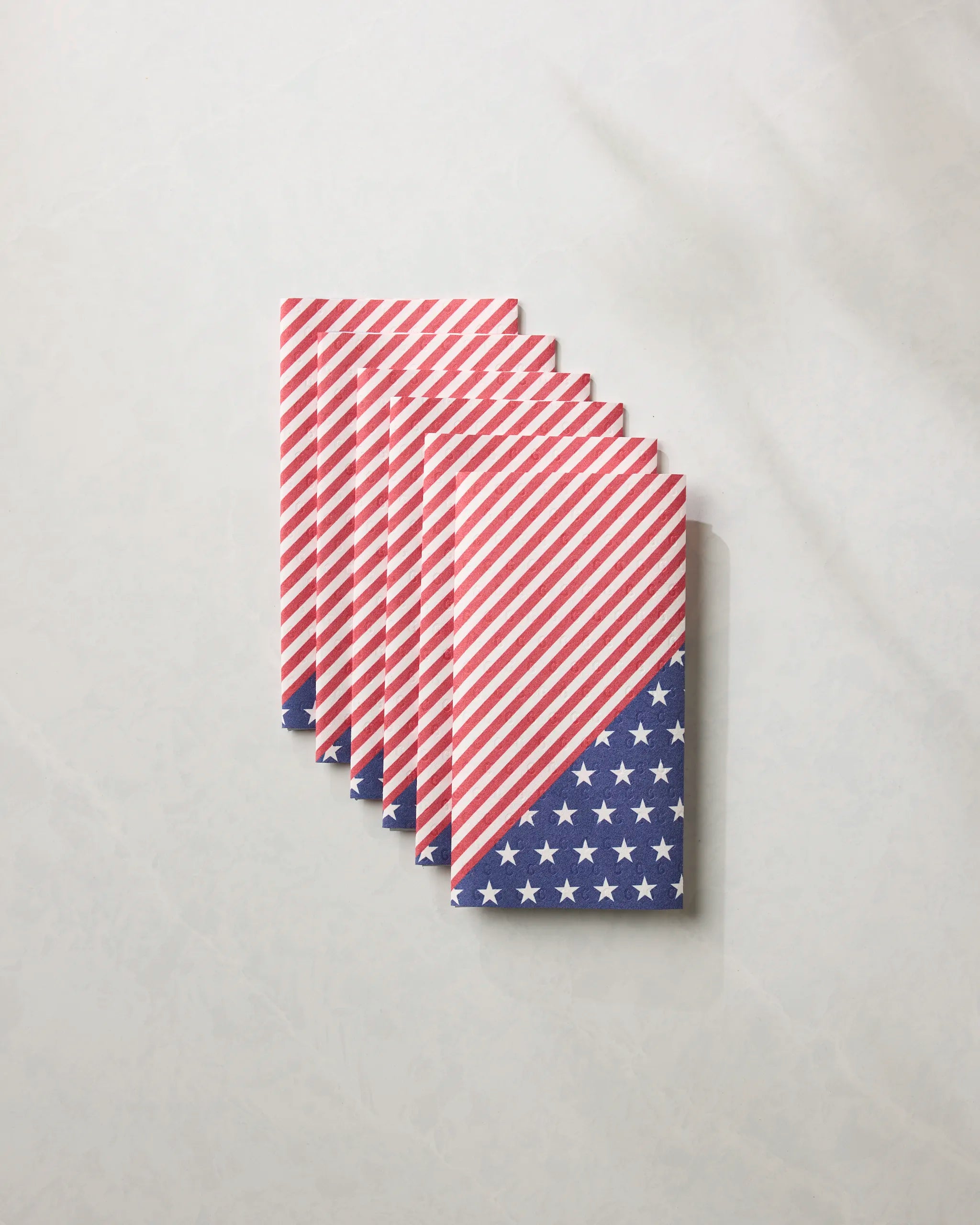 Six folded Geometry Liberty eco-friendly kitchen towels featuring an American flag-inspired pattern—red and white diagonal stripes with a blue corner of white stars—are neatly arranged in an overlapping row on a light surface.