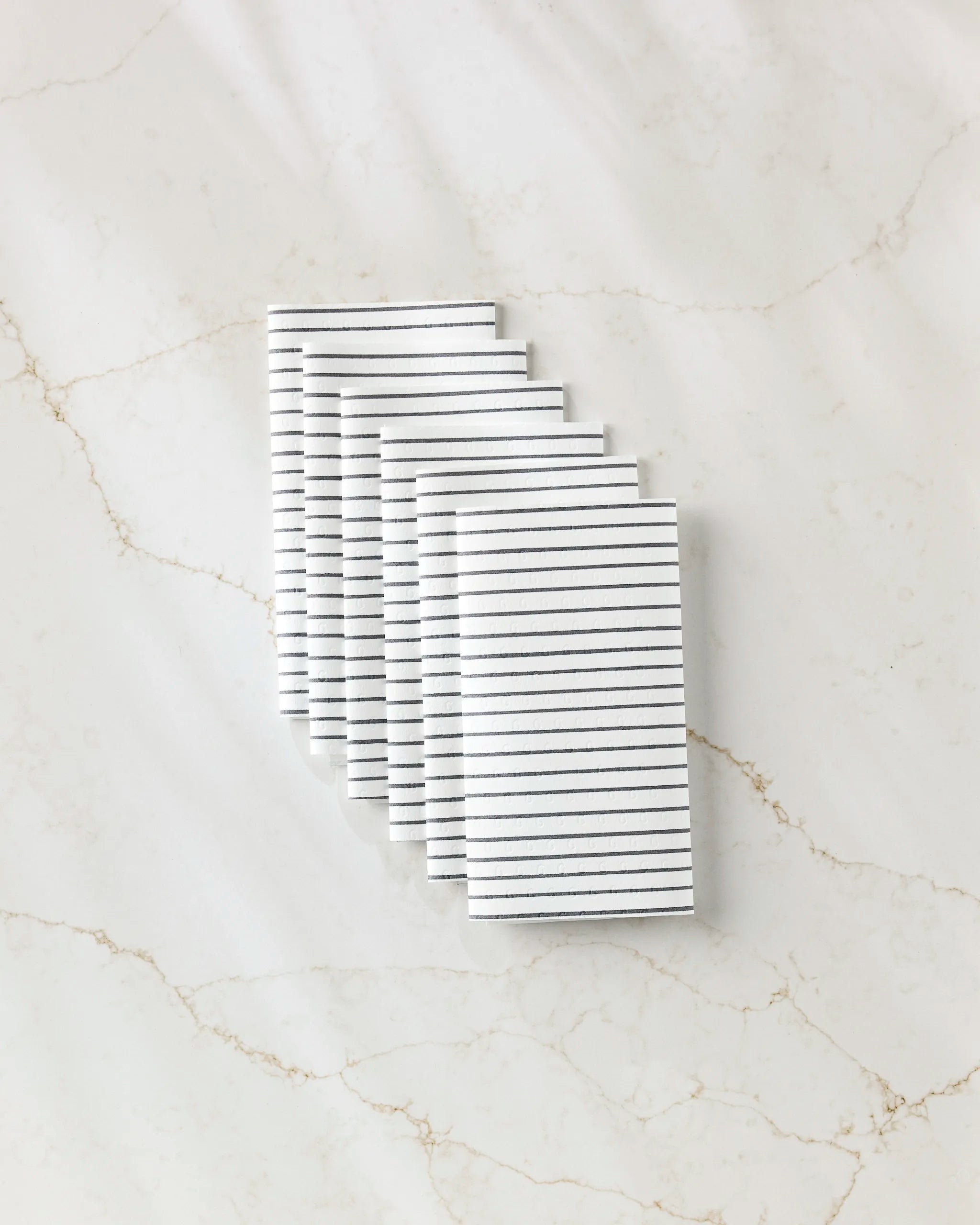 Seven GEOMETRY Linen white cloth napkins, each with thin black horizontal stripes and ideal as reusable paper towels, are neatly stacked on light brown-veined white marble.