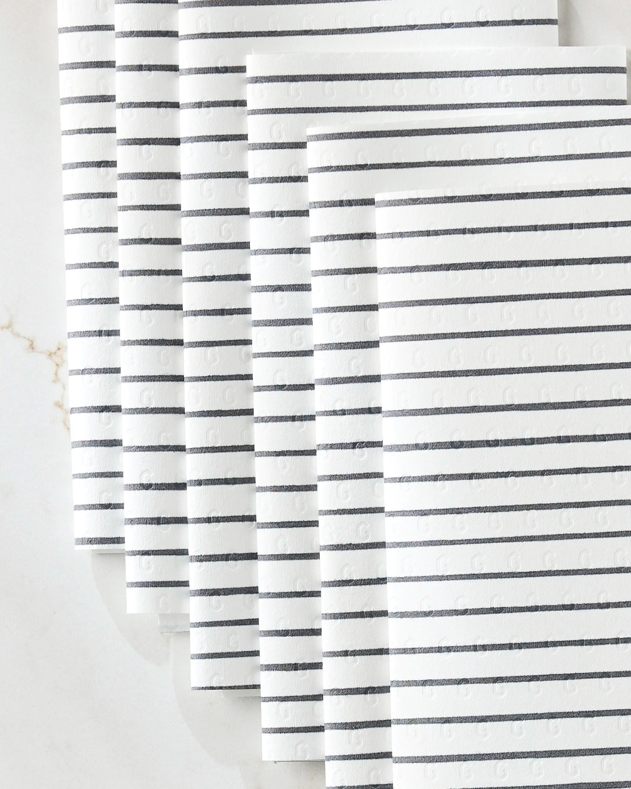 Six white eco-friendly "Linen" paper towels by GEOMETRY, featuring evenly spaced black horizontal lines, are neatly fanned out on a light marble surface.