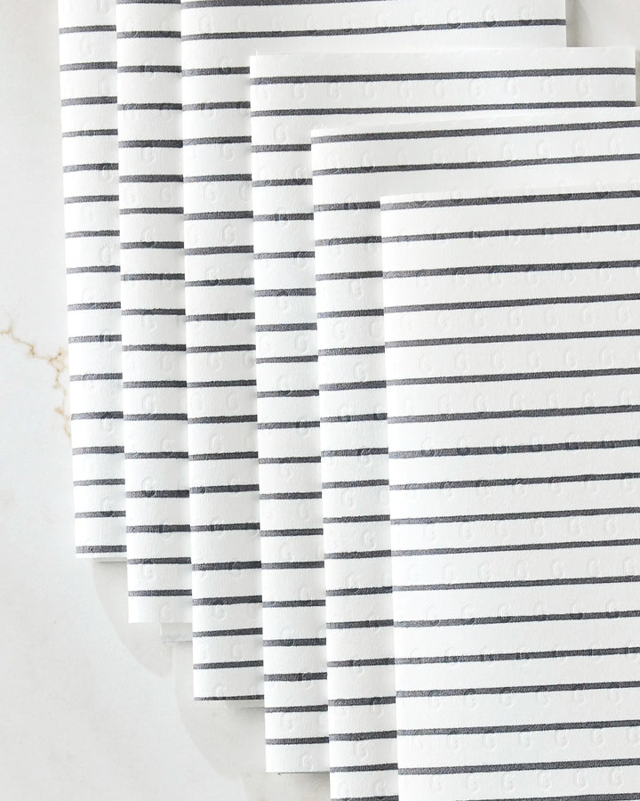 Six white eco-friendly "Linen" paper towels by GEOMETRY, featuring evenly spaced black horizontal lines, are neatly fanned out on a light marble surface.