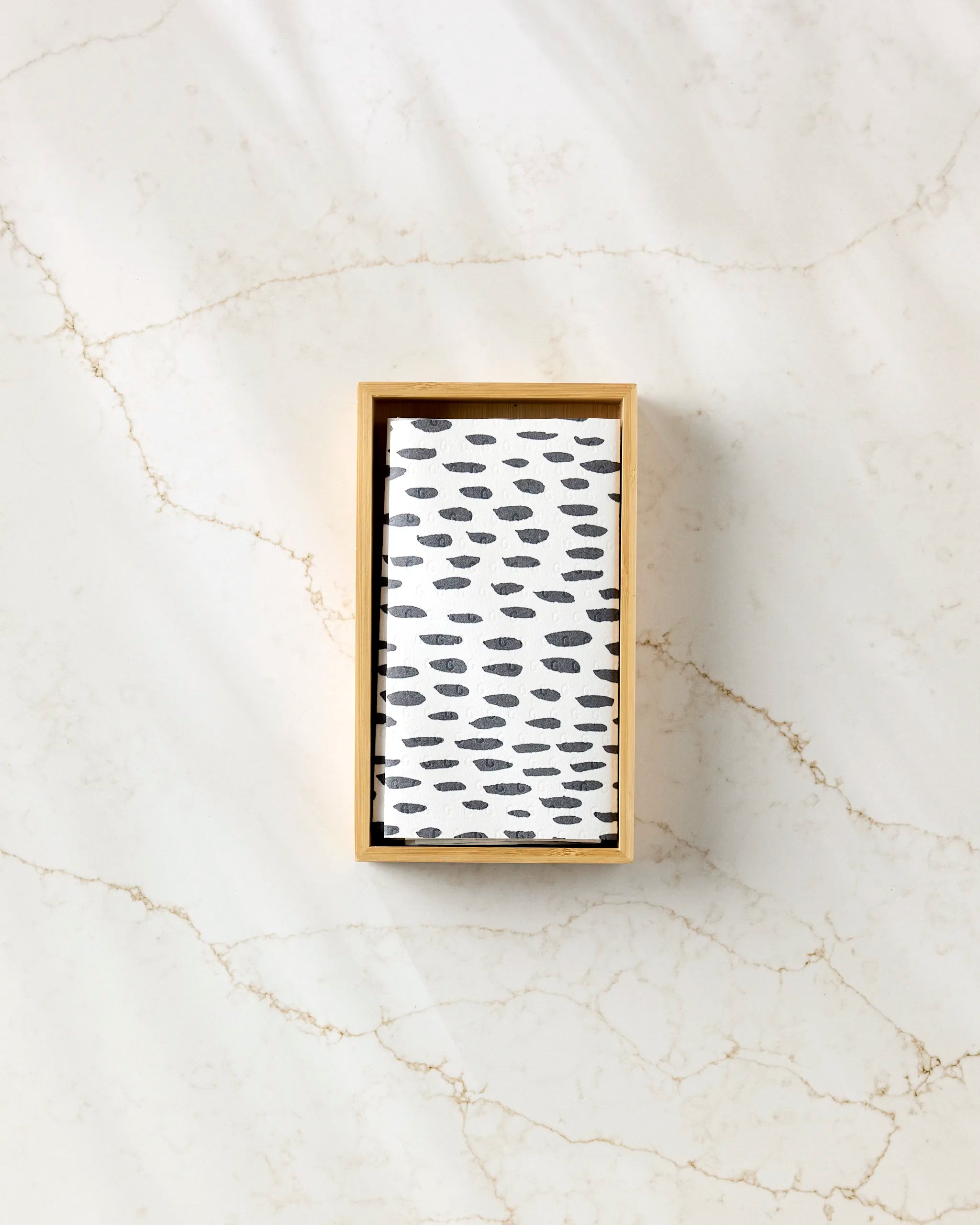 The GEOMETRY "Lines, Dots and Dashes" eco-friendly paper towel, featuring a black oval dot pattern, rests on a rectangular wooden tray set atop a subtly veined light marble surface.