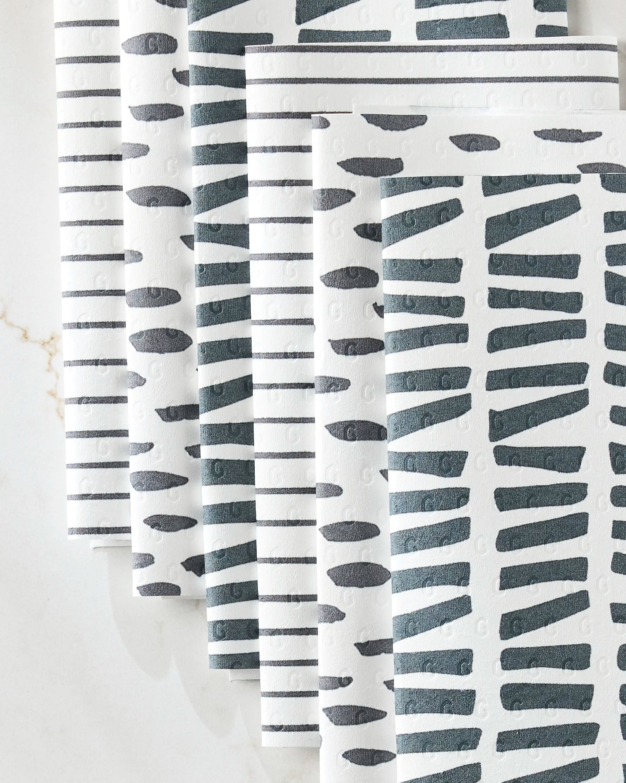 Several eco-friendly GEOMETRY "Lines, Dots and Dashes" paper towels are fanned out on a light surface, each sheet displaying bold dark gray geometric or striped designs with alternating vertical lines and horizontal brushstrokes.