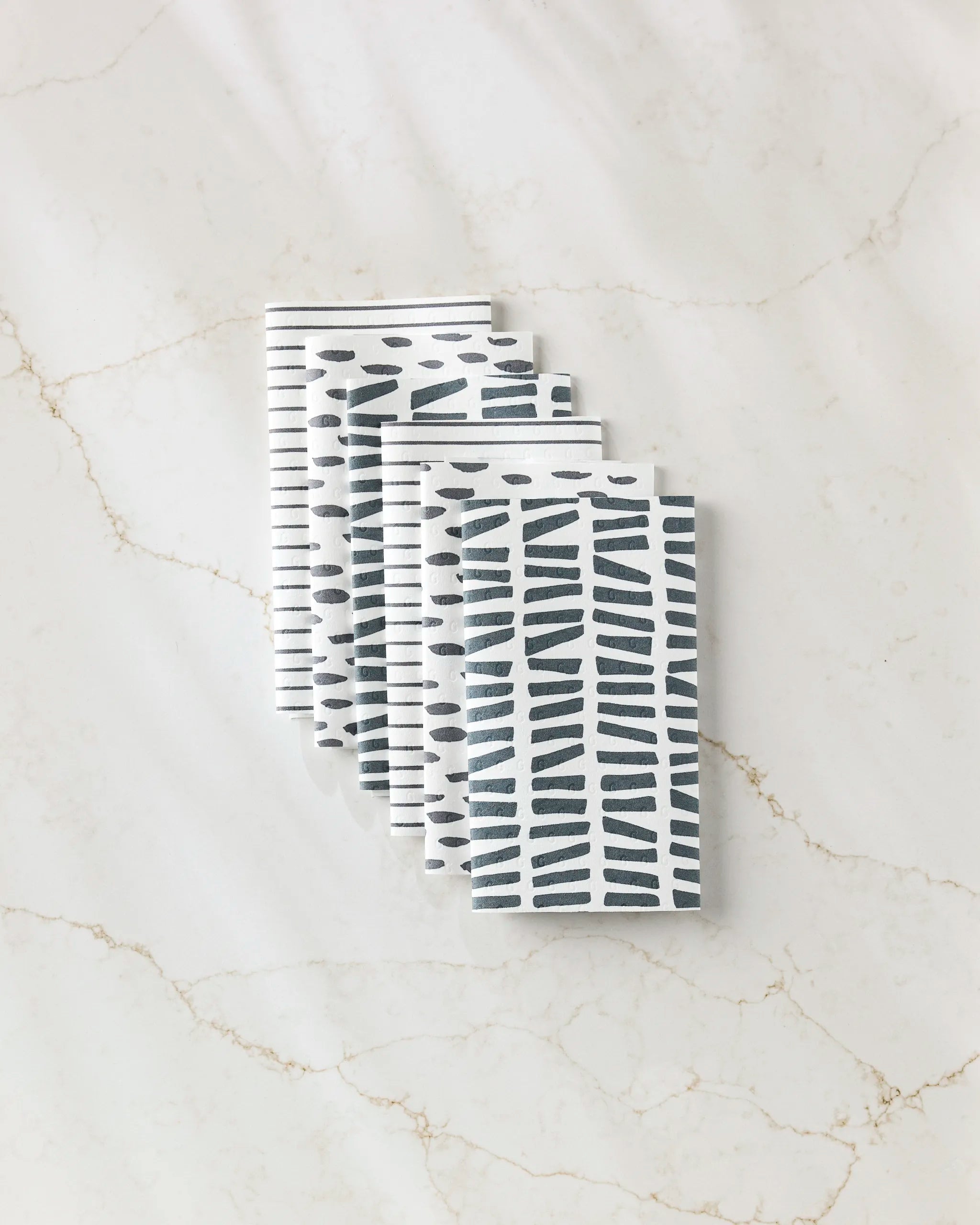 Four "Lines, Dots and Dashes" napkins by GEOMETRY, featuring black geometric patterns on white, are neatly stacked on a marble surface—an eco-friendly alternative to paper towels.