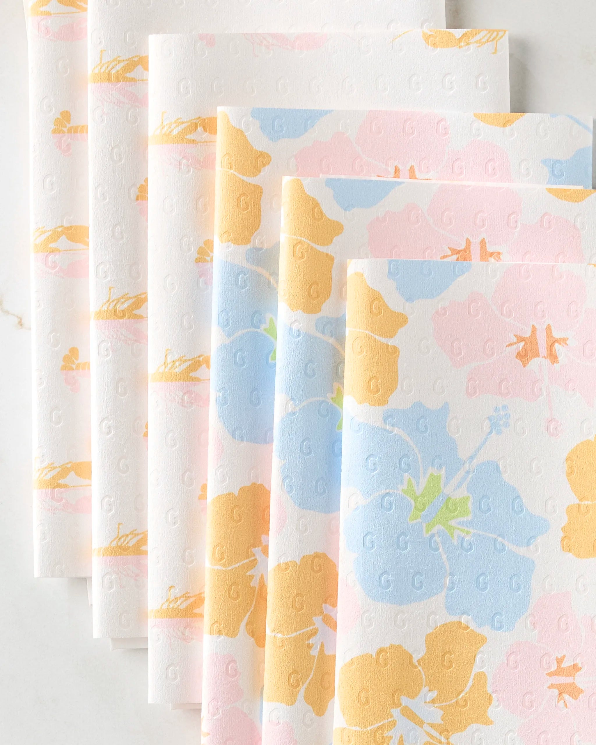 Several sheets of GEOMETRY’s Lobster Love paper towels, some plain white with small embossed Gs and others featuring a blue, pink, and yellow pastel floral design, are neatly arranged in an overlapping row.