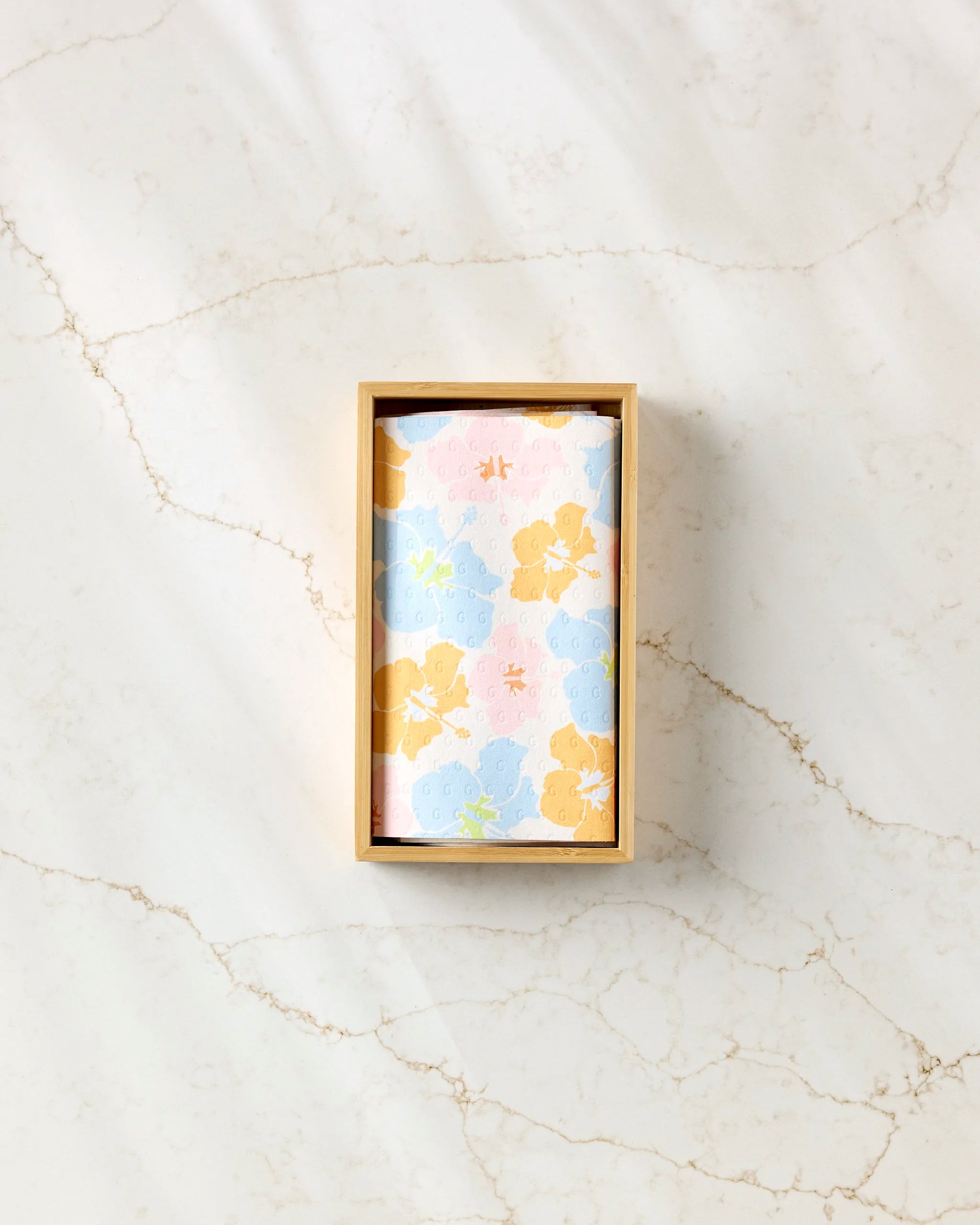 A rectangular wooden box from GEOMETRY holds "Lobster Love," a pastel floral eco-friendly cloth with blue, yellow, and pink flowers, displayed on a white marble surface streaked with light brown veins.