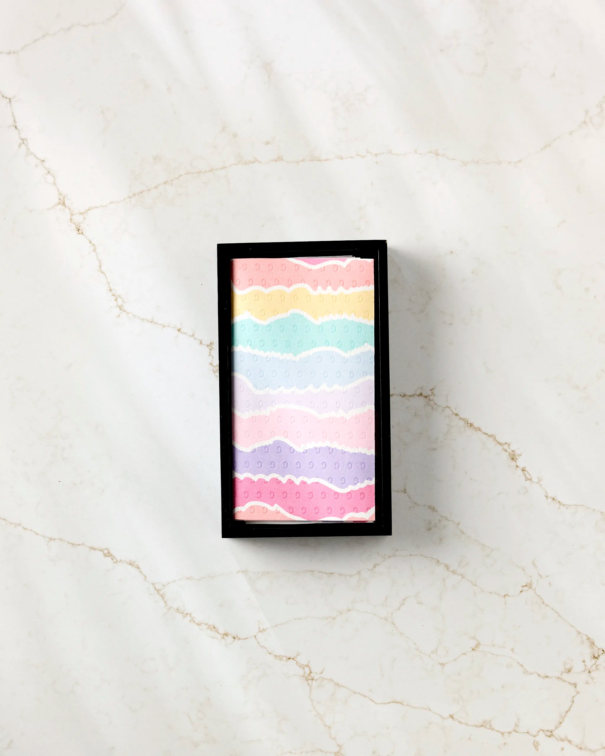 A rectangular black box from Rebecca Bobko holds the “Lots of Dots” eco-friendly paper towels with pastel stripes and wavy patterns, displayed on white marble with light brown veins.