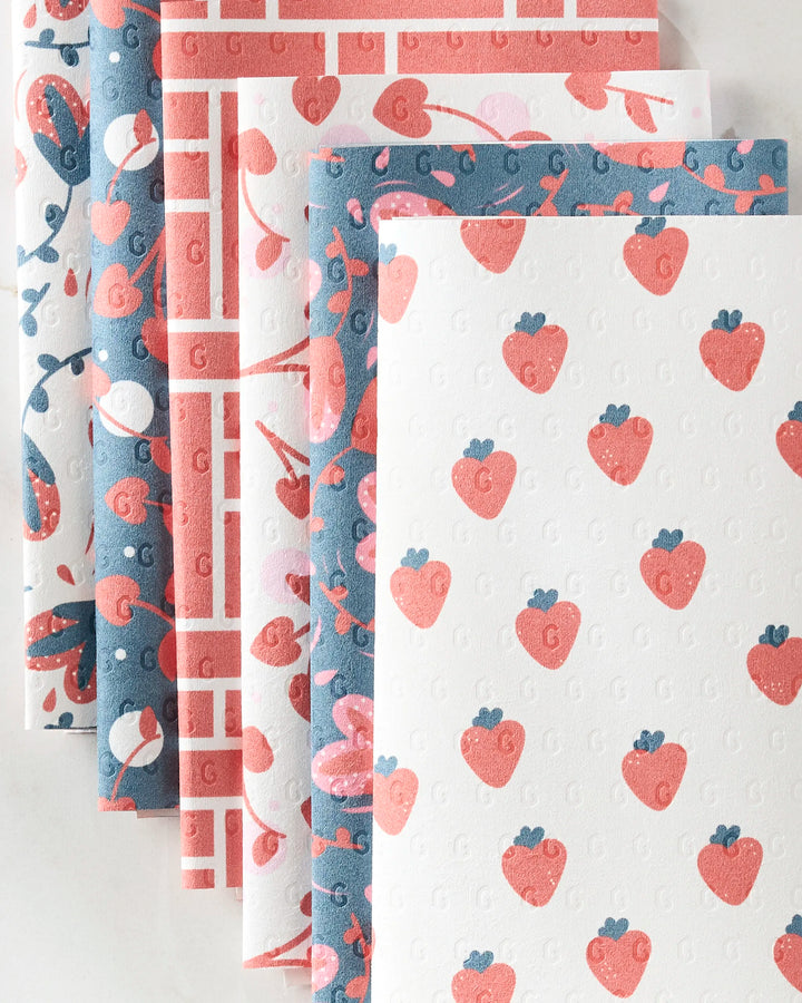 Six "Love Blooms" reusable paper towels by Charlie Rowan Designs, featuring pink, red, and blue strawberries, hearts, flowers, and geometric patterns, are fanned out as an eco-friendly kitchen towel alternative.