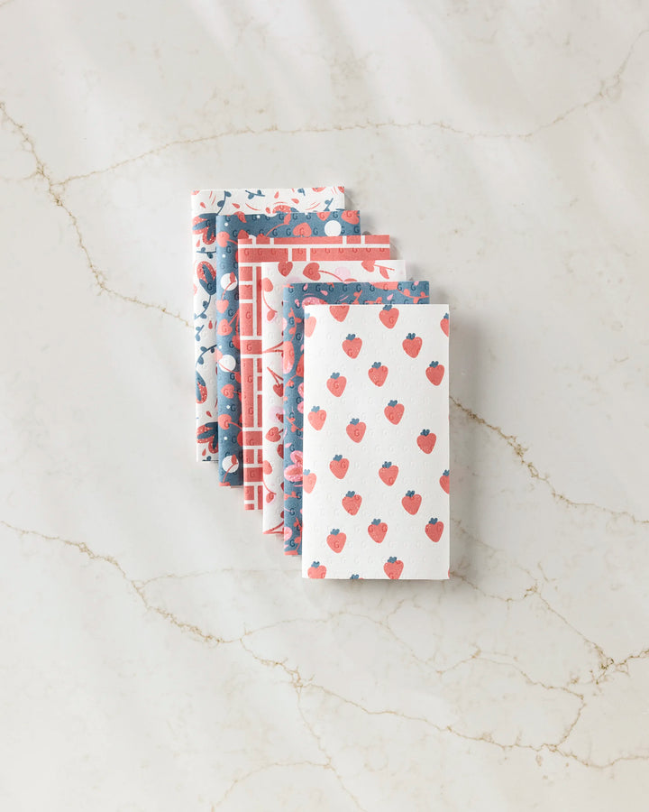 Love Blooms by Charlie Rowan Designs features four eco-friendly kitchen towels with blue and red strawberry and floral patterns, stacked on marble—a perfect reusable paper towel option for a sustainable home.