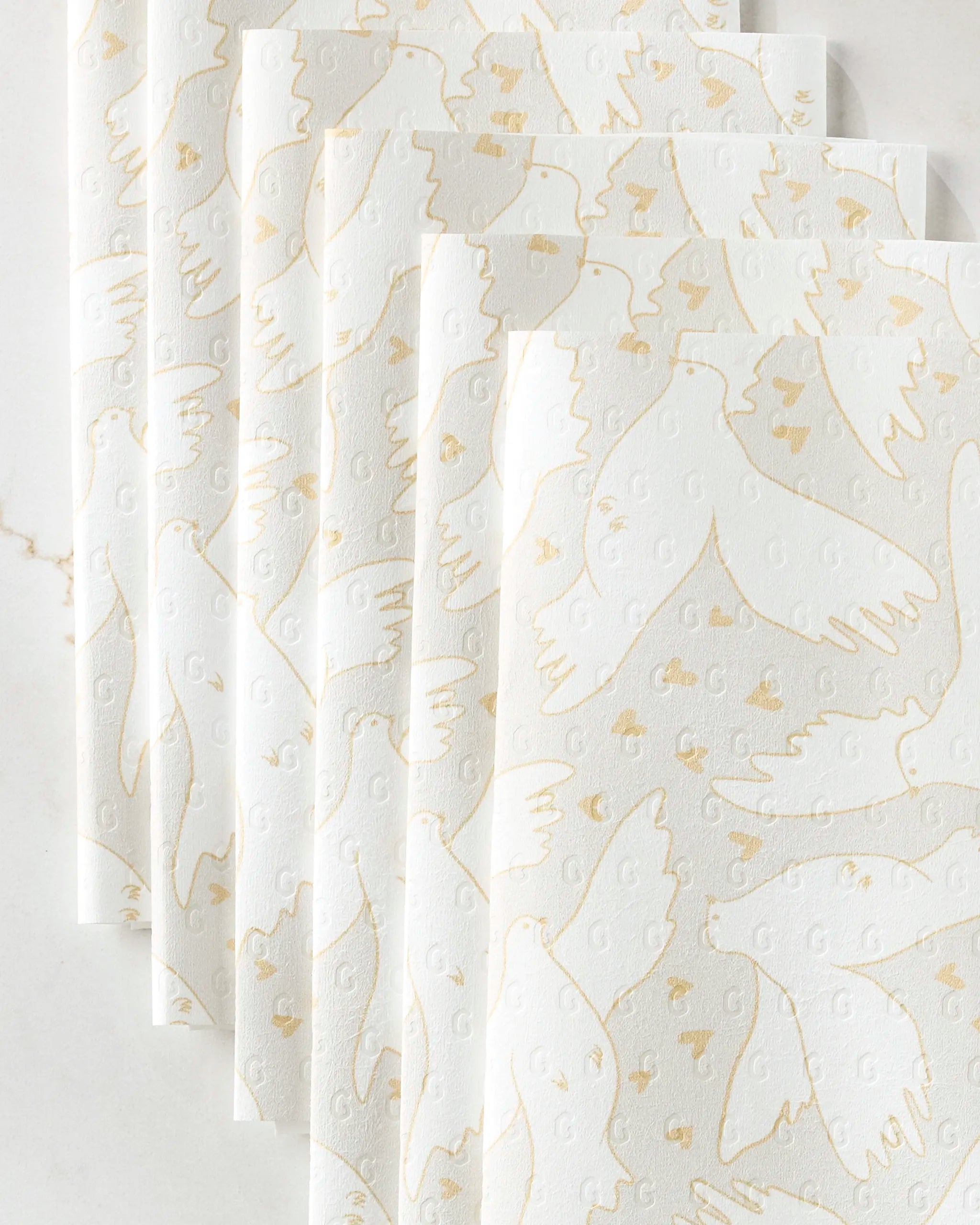 GEOMETRY’s "Love Takes Flight" white paper napkins, designed to look like eco-friendly towels, feature gold-outlined flying doves and hearts on a textured surface, beautifully displayed on a white marble background.
