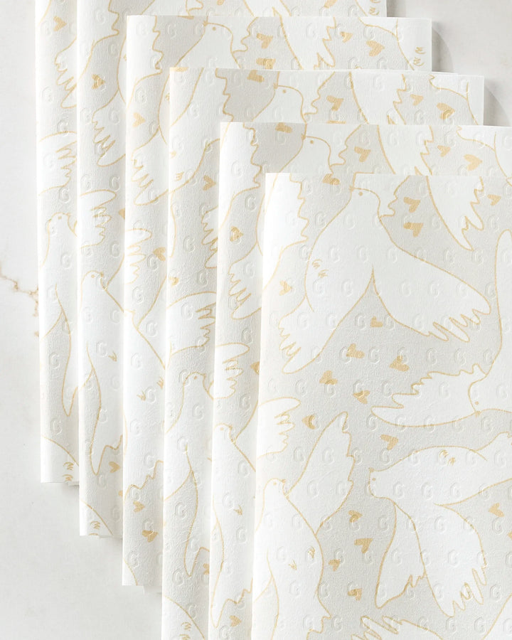 GEOMETRY’s "Love Takes Flight" white paper napkins, designed to look like eco-friendly towels, feature gold-outlined flying doves and hearts on a textured surface, beautifully displayed on a white marble background.