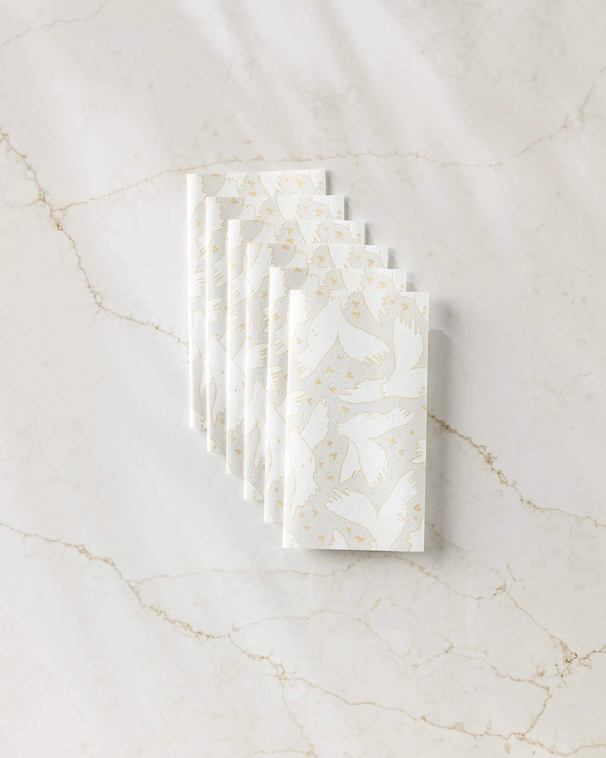 Six folded, reusable GEOMETRY "Love Takes Flight" paper towels, featuring a subtle light beige bird and branch pattern, are neatly arranged in a row on white marble with light brown veining.