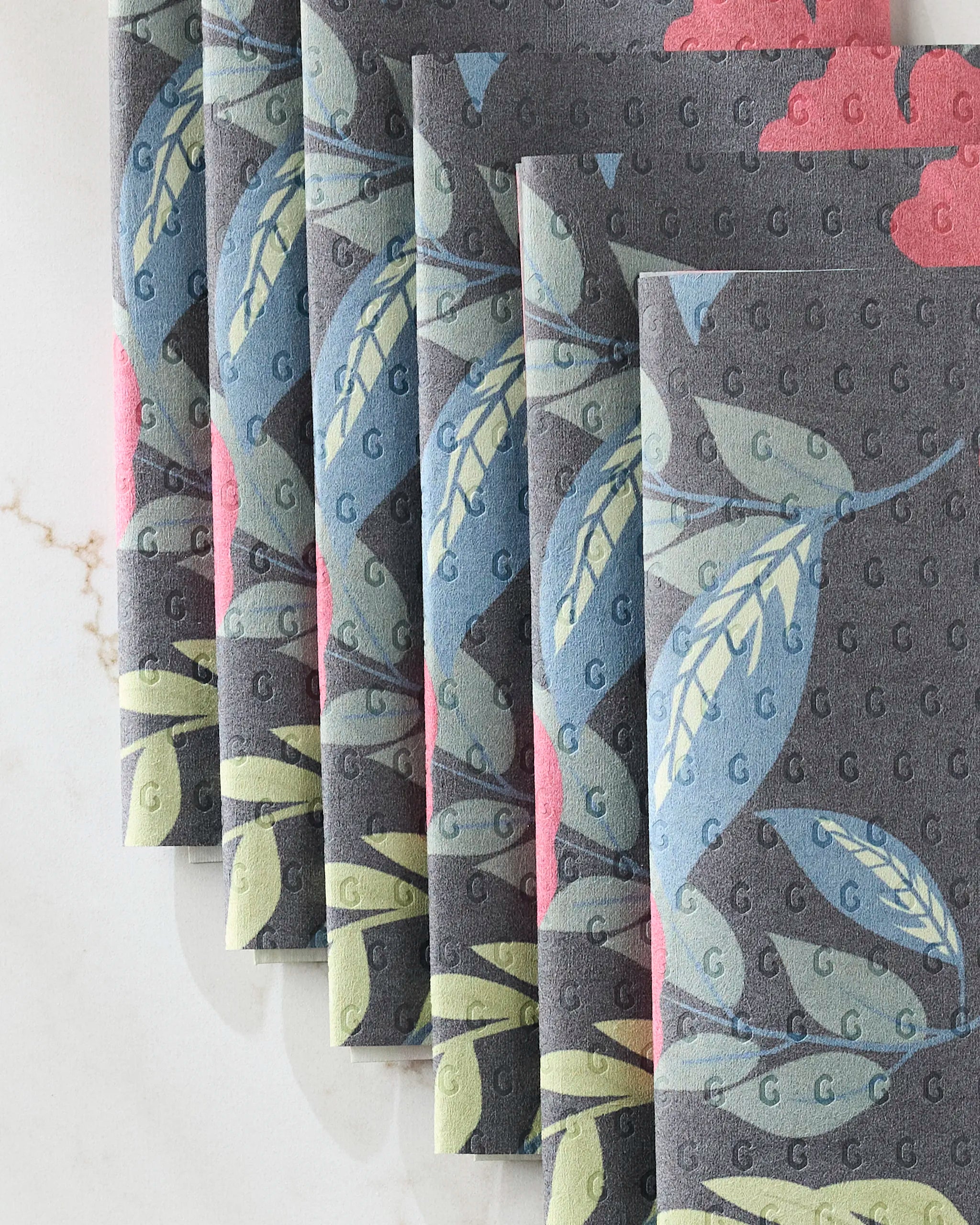 Lovely Night napkins by GEOMETRY feature a gray base with blue and green leaves, pink accents, and repeating small G patterns. These eco-friendly paper towels are neatly overlapped on a light marble surface.