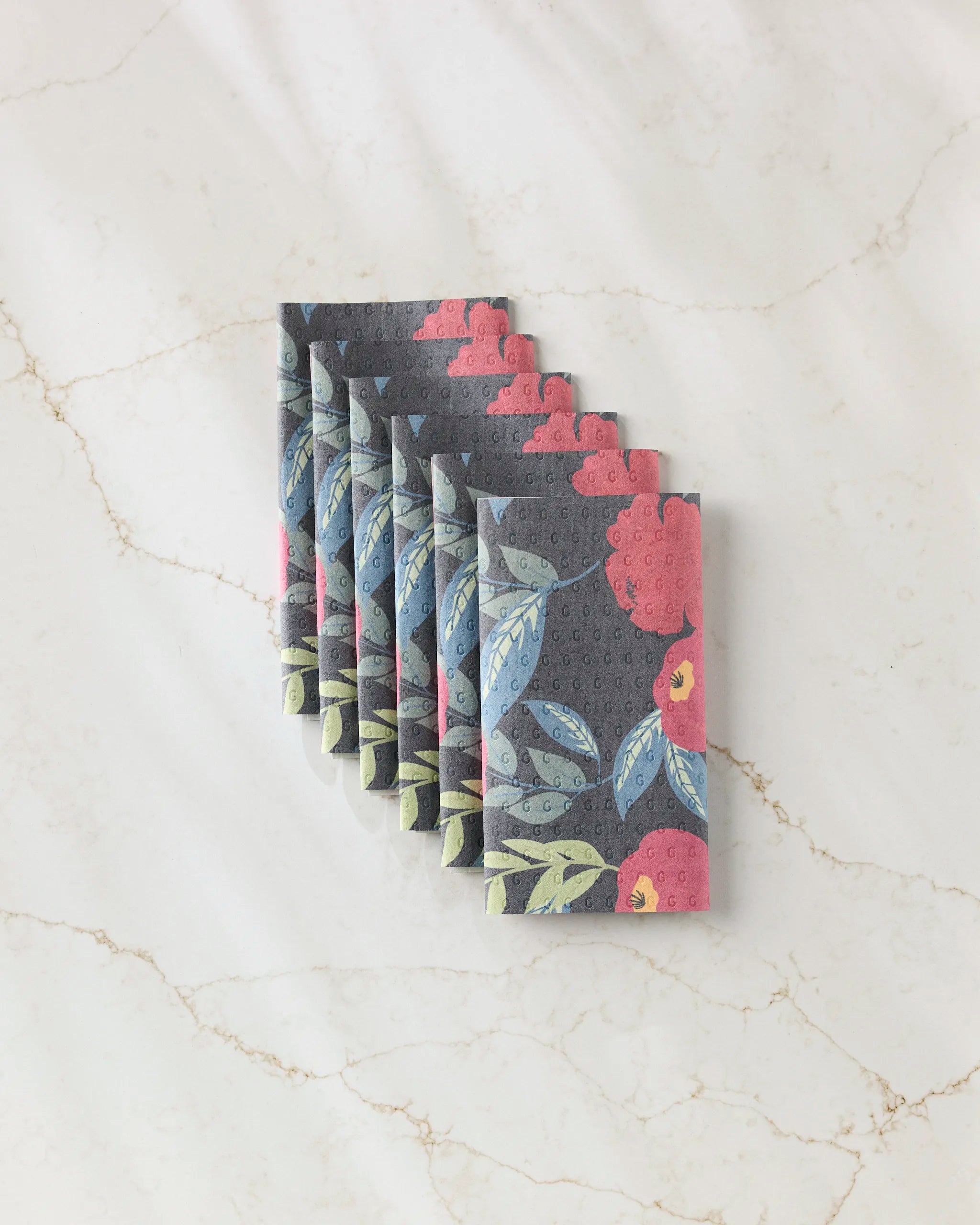 Six folded GEOMETRY "Lovely Night" cloth napkins, featuring a floral pattern in pink, blue, green, and gray tones, are elegantly fanned on white marble—a stylish and eco-friendly alternative to disposable paper towels.