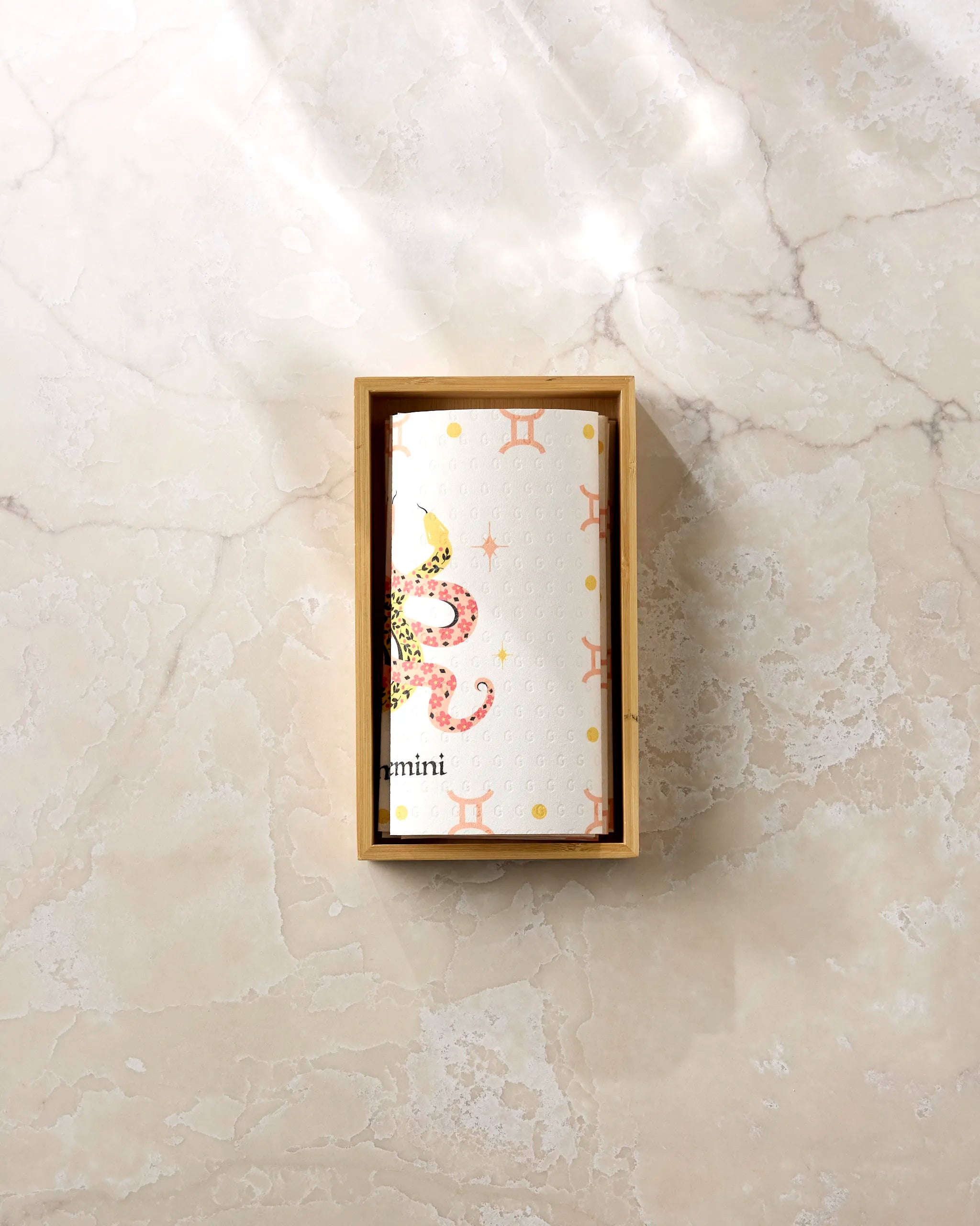 The Lucky Gemini by Heather Zhou is a white cylindrical item with Gemini-inspired yellow stars and orange zodiac motifs, set in a wooden box on marble—crafted from eco-friendly, post-consumer recycled paper towels.