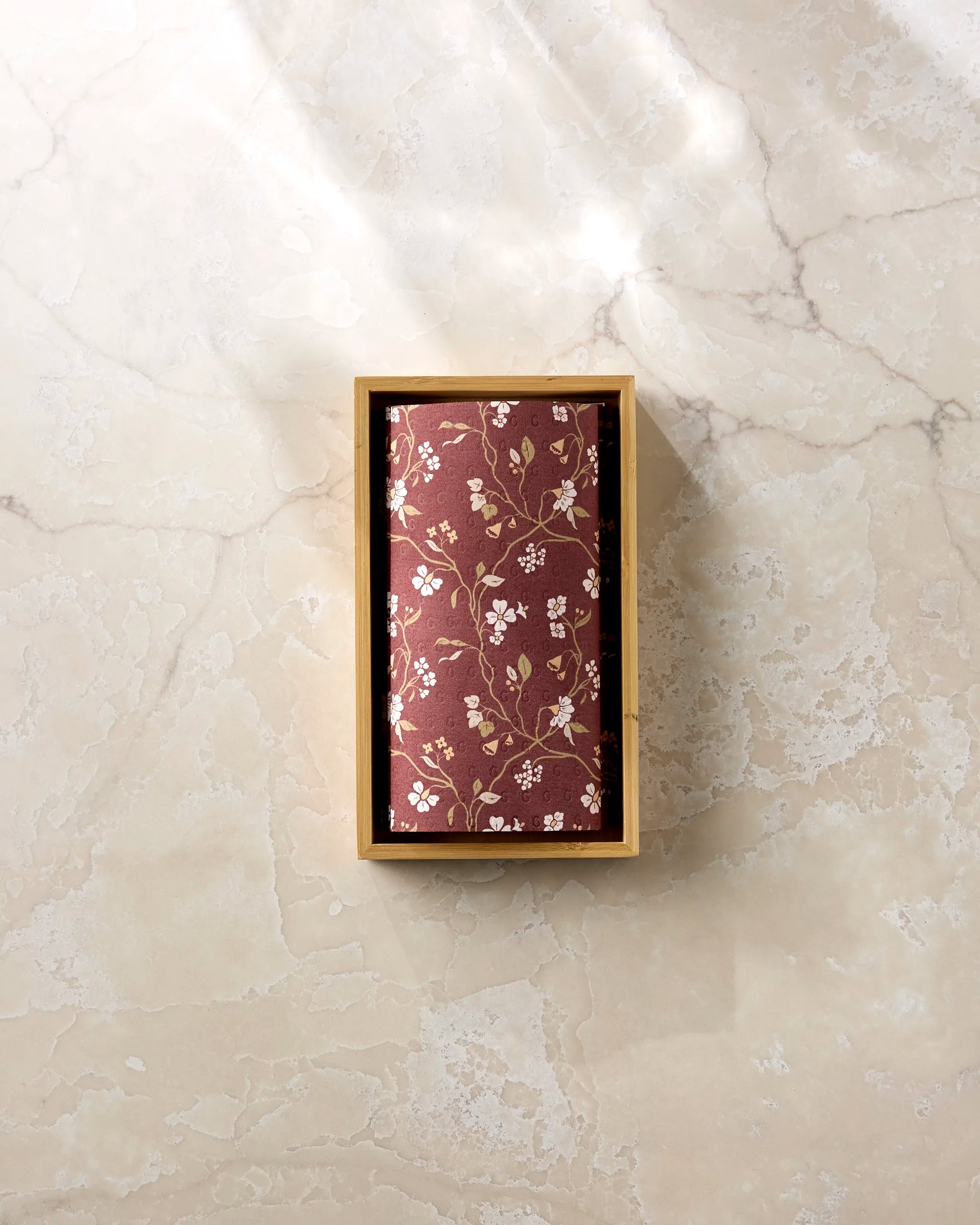 A rectangular box of Lunaria in Merlot by Danelys Sidron, featuring a red floral pattern and made from post-consumer recycled materials, sits on a light marble surface bathed in gentle sunlight.