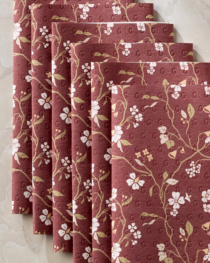 Five Lunaria in Merlot napkins by Danelys Sidron, featuring white flowers and green leaves on muted red, are neatly stacked on marble—an eco-friendly alternative to disposables.