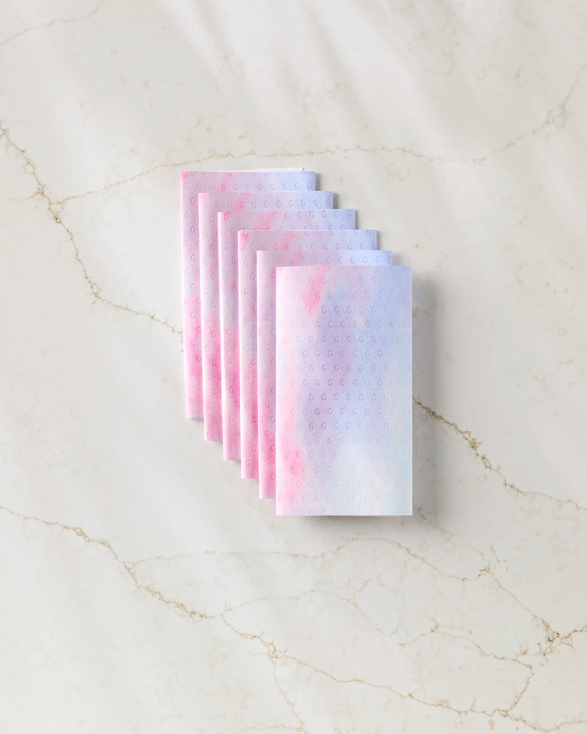 Seven pastel-colored "Magic In The Sky" cloths by Stefanie Bales, featuring a pink and blue gradient, are neatly stacked on a marble surface as an eco-friendly, stylish alternative to paper towels for your kitchen.