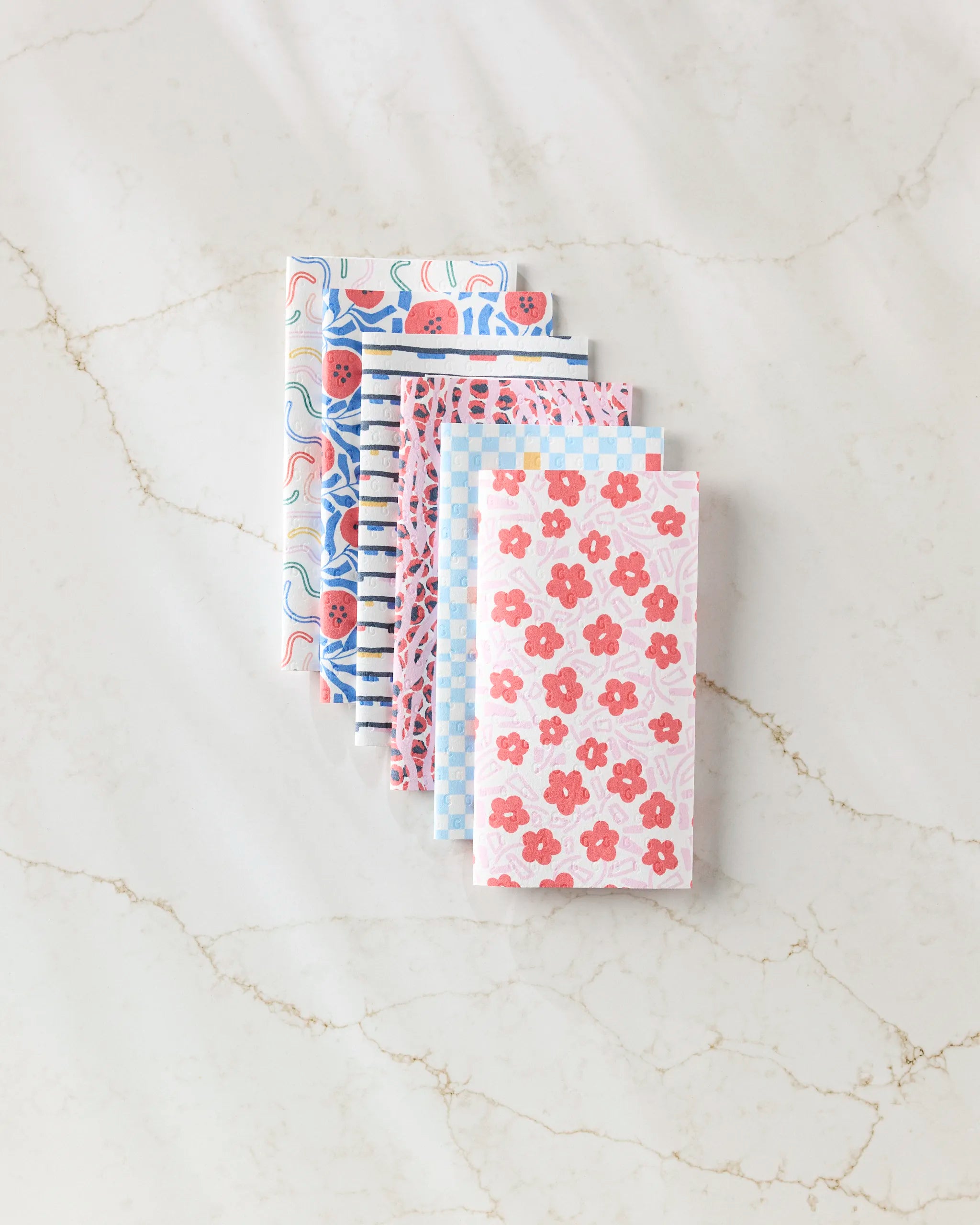 Five folded Magic Wand reusable paper towels by Happy Hemera, featuring assorted floral and striped patterns in pink, red, and blue hues. Arranged fan-shaped on a light marble surface for an eco-friendly display.