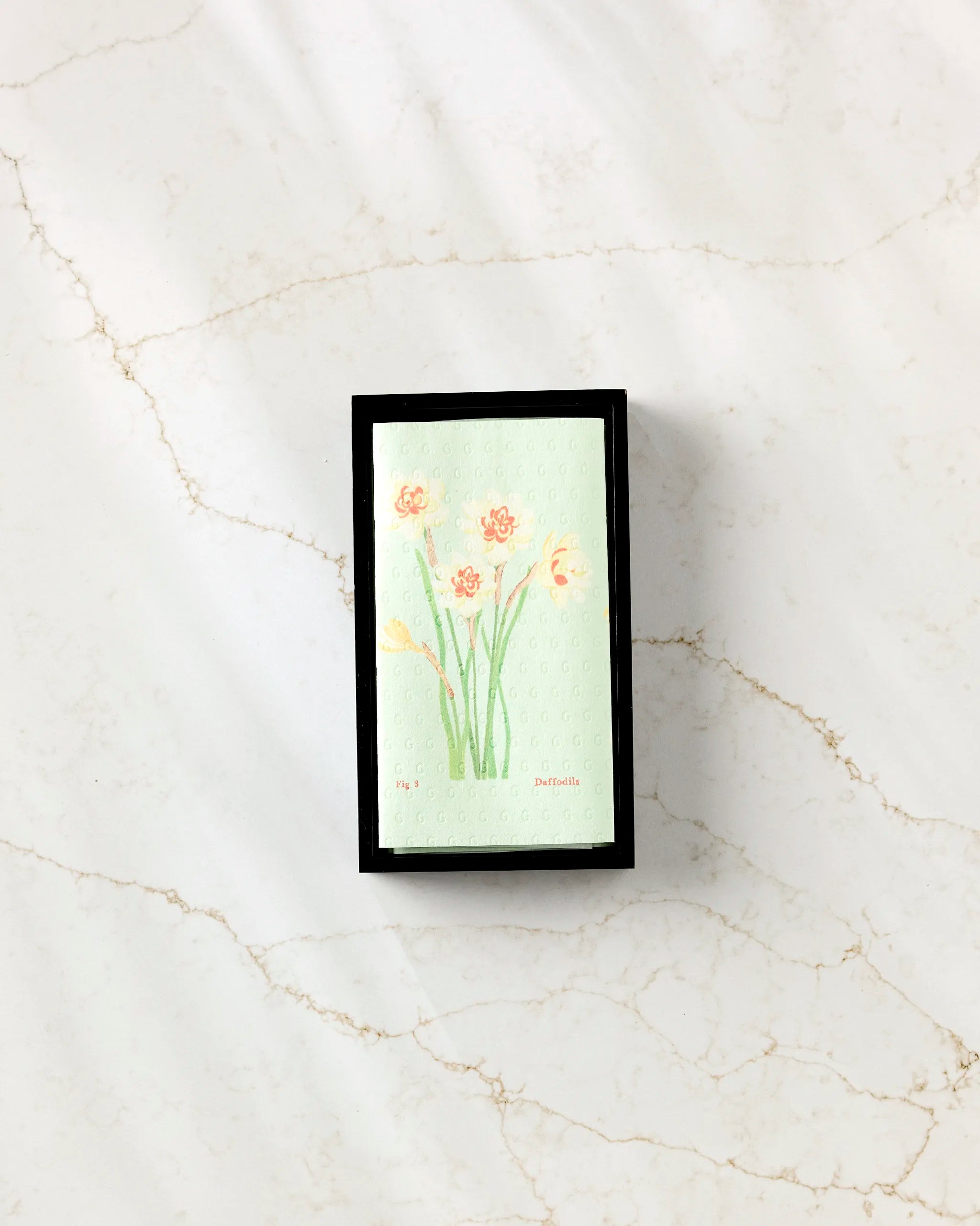The “March Daffodils” card from Linda Fox features vibrant orange and yellow daffodils on a light green background with a black border, centered on marble to evoke the fresh, eco-friendly spirit of spring.