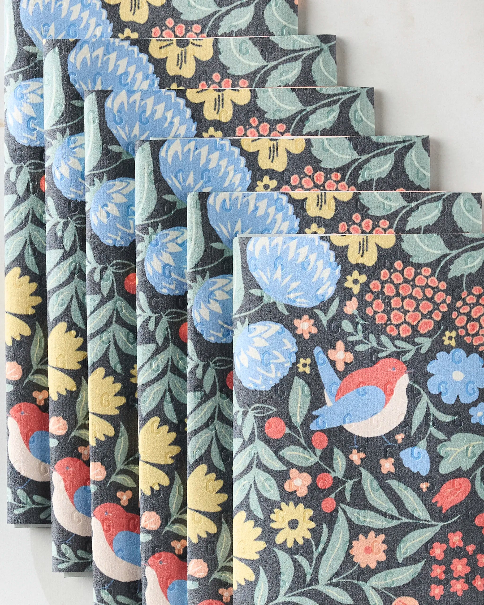 Six “Marsh Wren” cloth napkins by Danelys Sidron, featuring a repeating botanical pattern with blue flowers, yellow accents, red berries, green leaves, and a red and blue bird on dark background—an eco-friendly paper towel alternative.