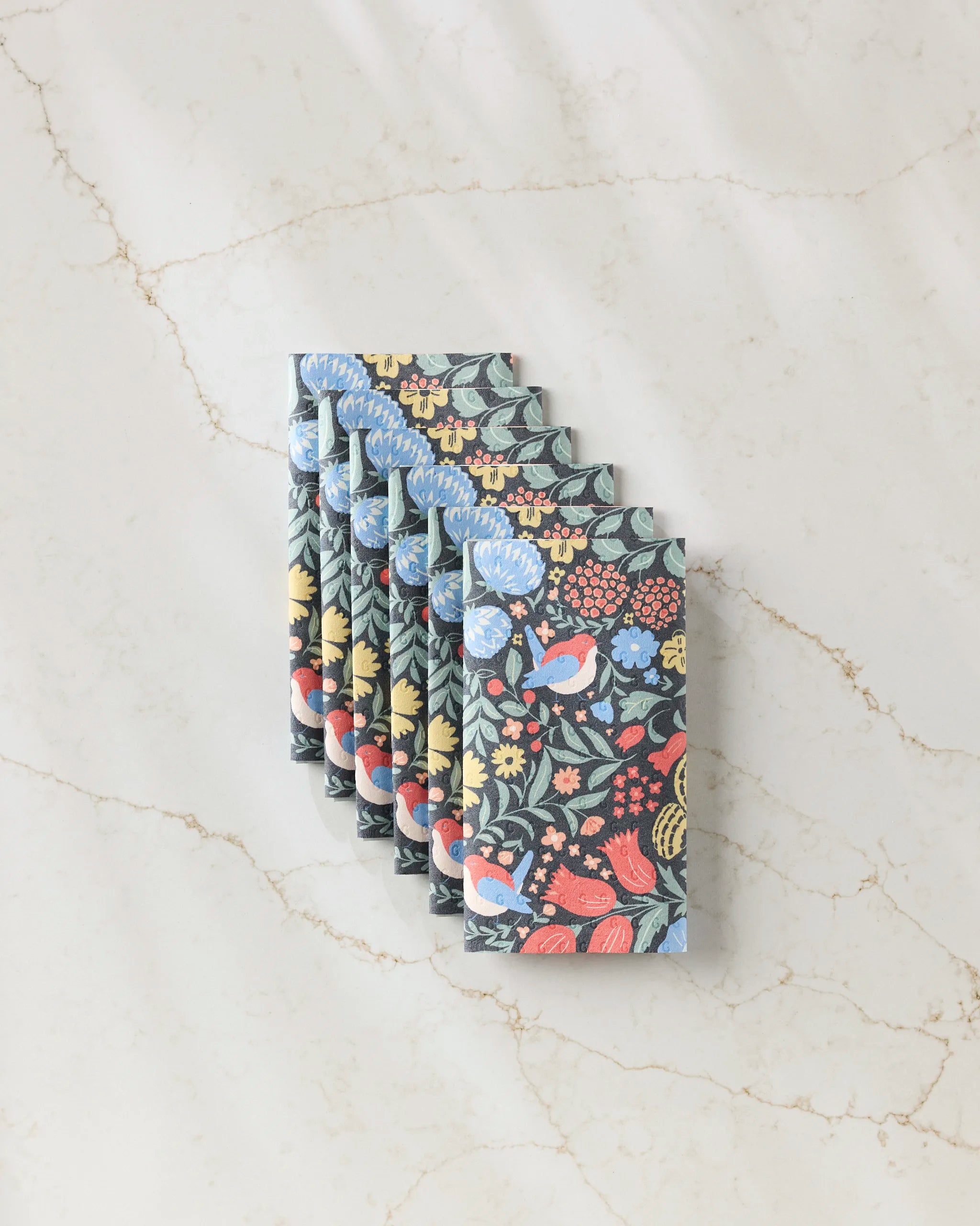 Six Marsh Wren reusable paper towels by Danelys Sidron, featuring a colorful floral and bird pattern, are neatly arranged in an overlapping stack on a light marble surface.