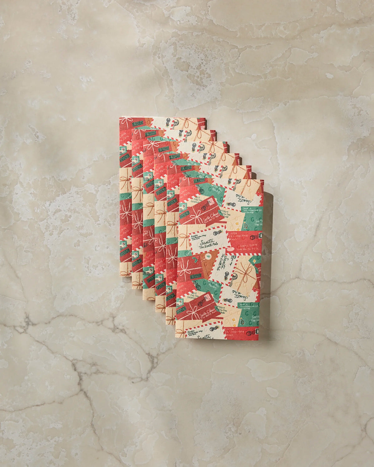 Seven folded Geometry “Merry Mail” holiday-themed reusable paper towels, featuring red, green, and beige gift and ribbon designs, arranged in a fanned-out stack on a light marble surface—an eco-friendly alternative to disposable paper towels that’s perfect for festive gatherings.