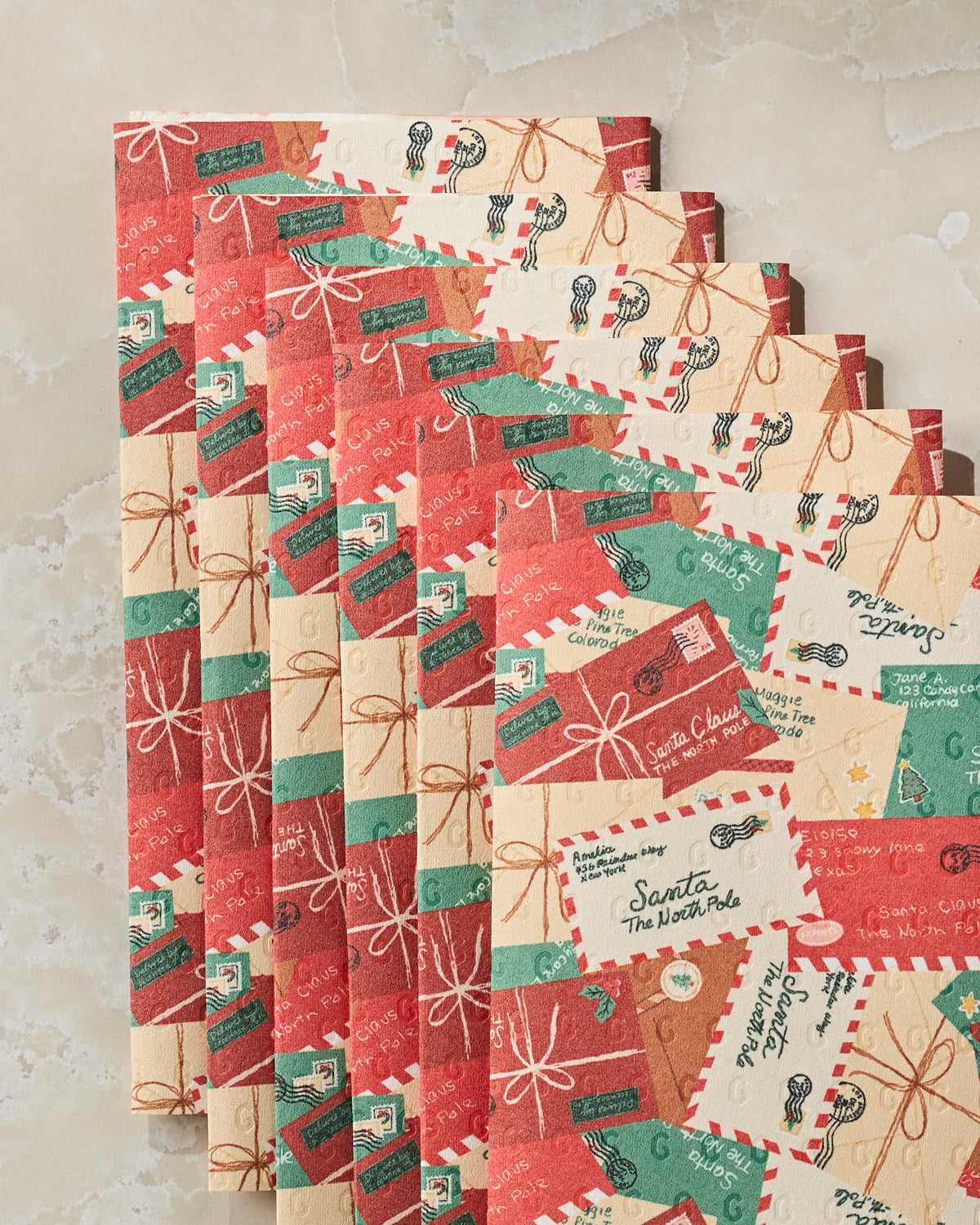 Geometry’s Merry Mail features six sheets of holiday-themed wrapping paper, adorned with charming stamps, postal markings, and festive hues of red, green, and cream. Arranged in a fanned-out stack on a light-colored surface, these wraps offer an eco-friendly alternative to disposable paper towels—bringing both sustainability and festive charm to your gift giving this holiday season.