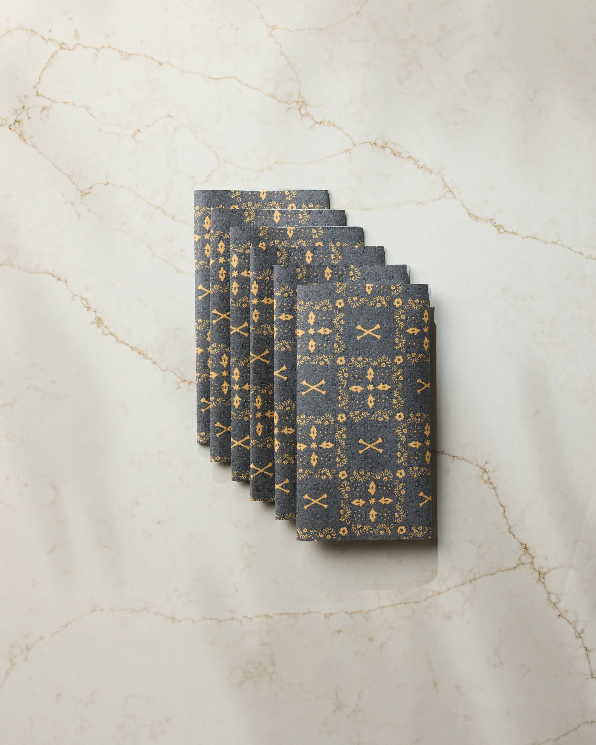 A stack of Geometry’s Midnight Bandana reusable towels, featuring a gold floral and geometric design, is neatly arranged on a white marble surface with tan veins.