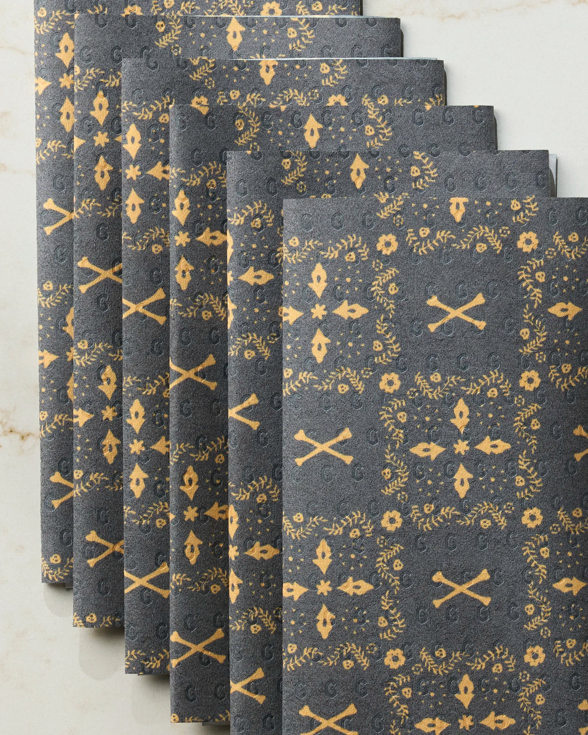 Six Midnight Bandana reusable paper towels by Geometry, featuring a gold floral and crossbones pattern on dark grey, are neatly stacked on a light marble surface.