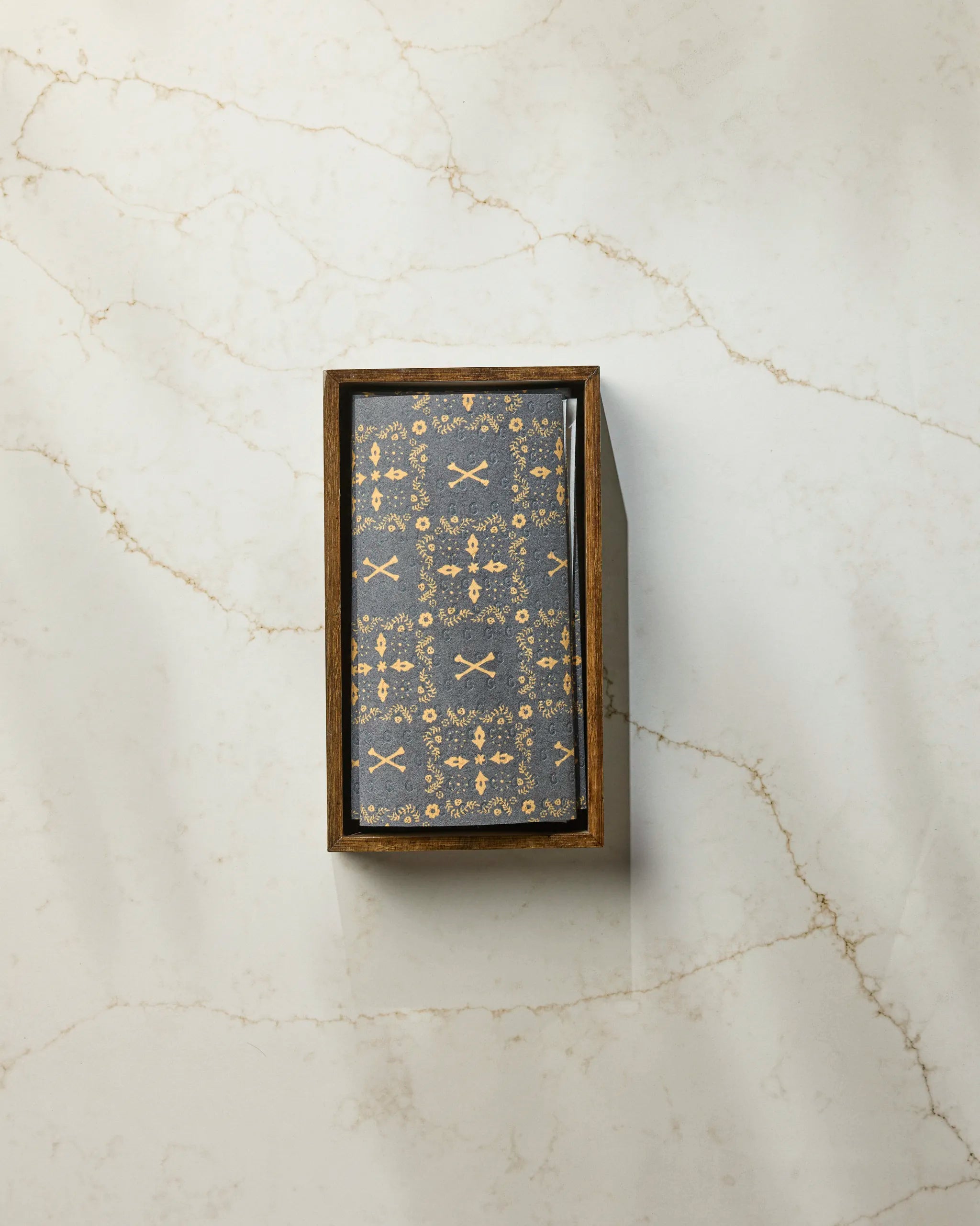 A box of Geometry's Midnight Bandana playing cards with an ornate gold and dark blue design sits on a white marble surface with light brown veins, next to a roll of eco-friendly paper towels.