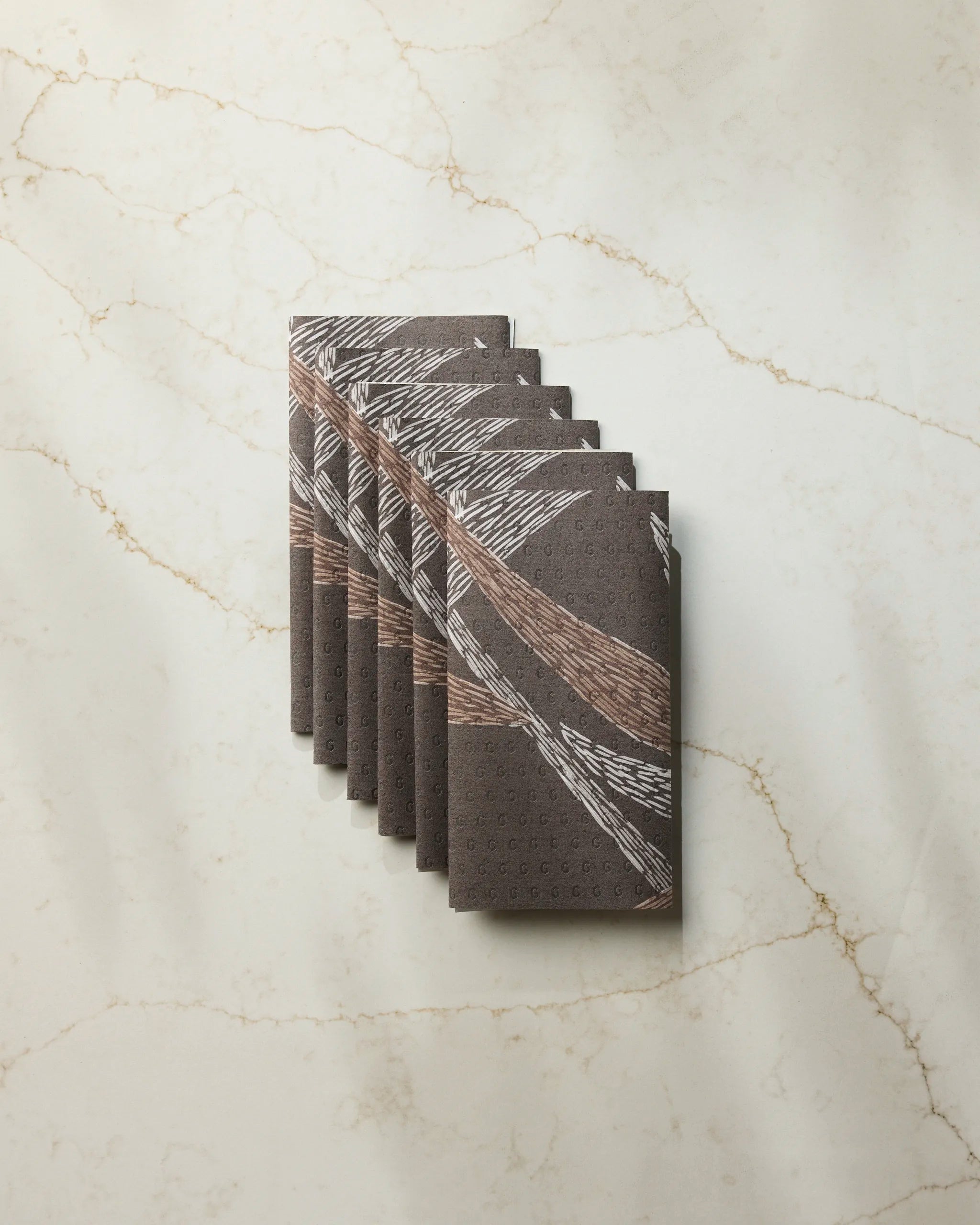Six folded Midnight Wildflower reusable paper towels by Geometry, featuring a white and tan abstract leaf pattern, are neatly fanned out on a light marble surface with subtle beige veining.