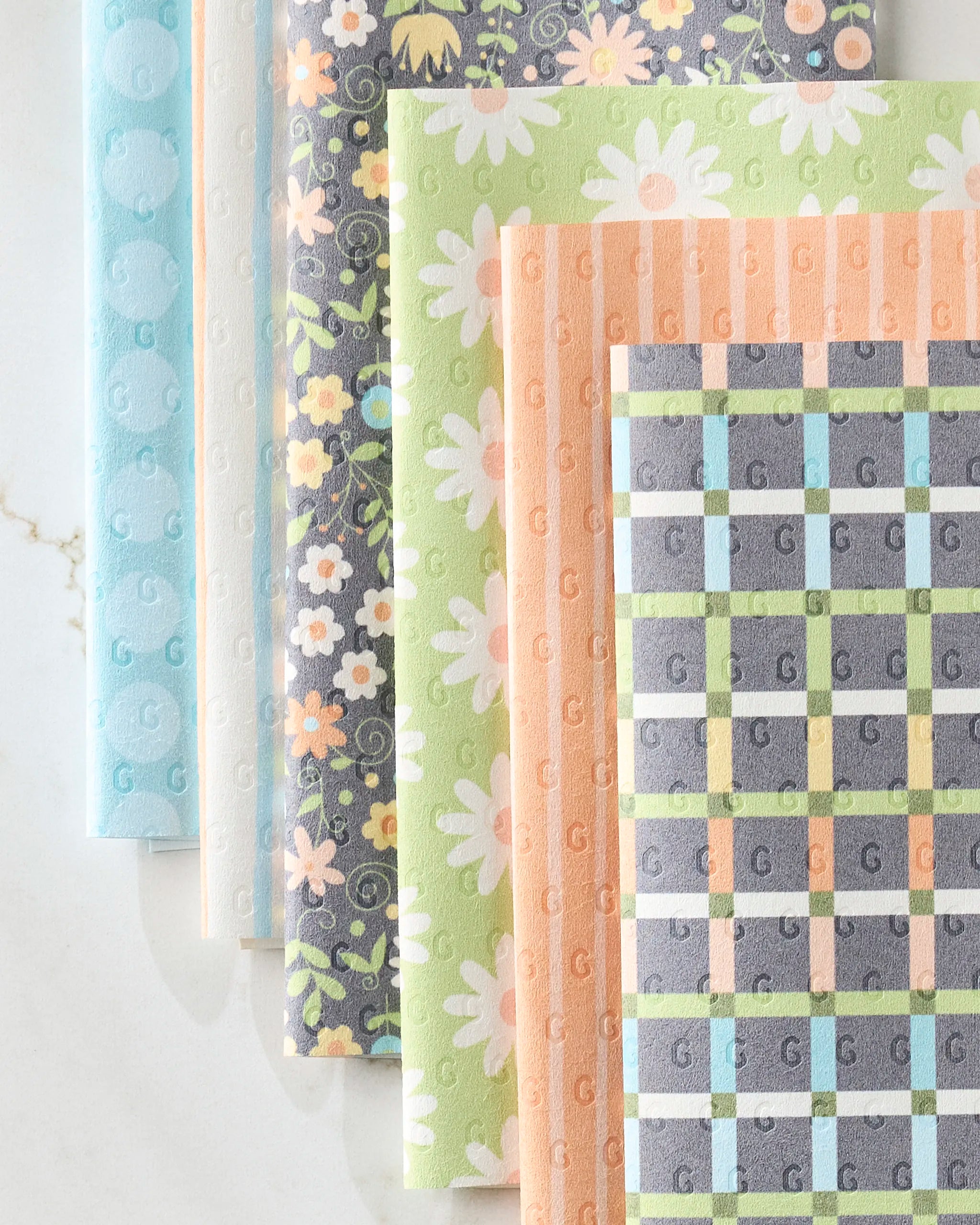 Five patterned Mindful Medley kitchen towels by Mindfulnice are displayed side by side on a light marble surface, featuring blue dots, pastel flowers, green daisies, dark blue floral, and colorful plaid designs.